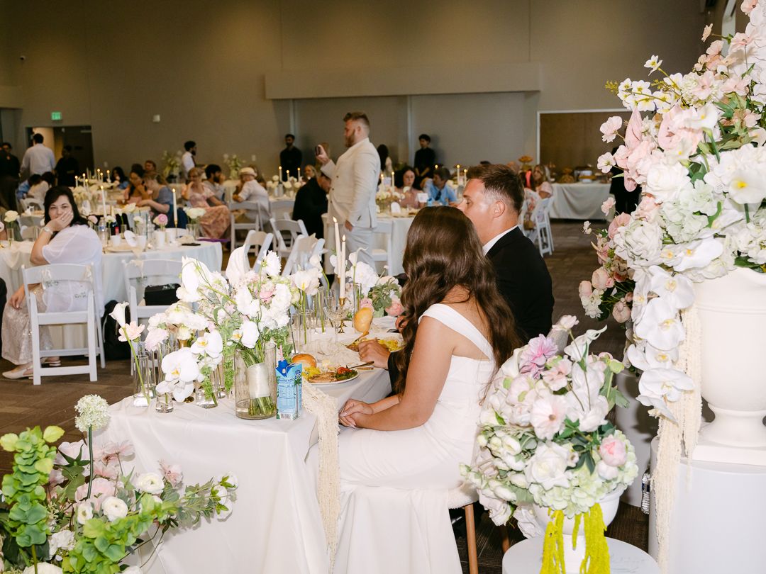 Bride and groom sit at sweetheart table surrounded by white floral centerpieces at elegant reception.