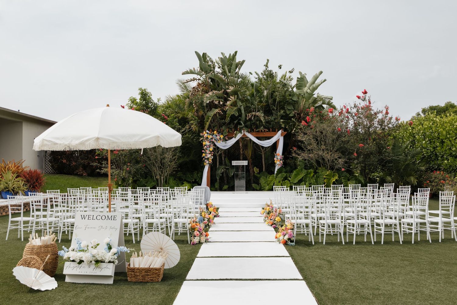 Elegant outdoor wedding ceremony setup with white chairs, floral arch, white aisle runner, and garden backdrop.