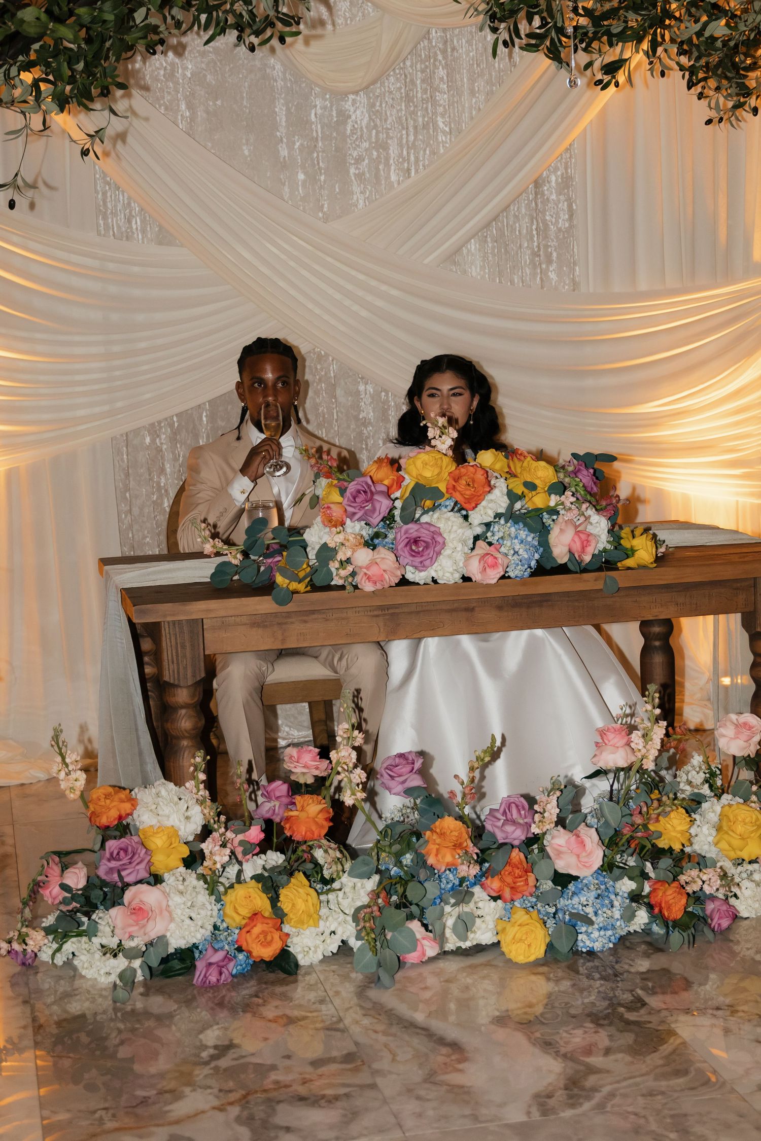 Bride and groom sitting at floral-adorned sweetheart table with colorful blooms under elegant draped backdrop.