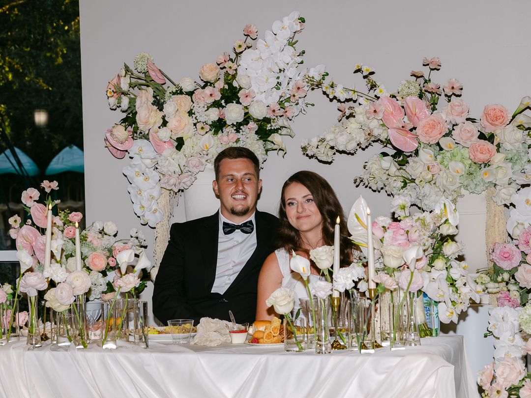 Newlyweds seated at elegantly decorated sweetheart table with pink and white floral backdrop at reception.