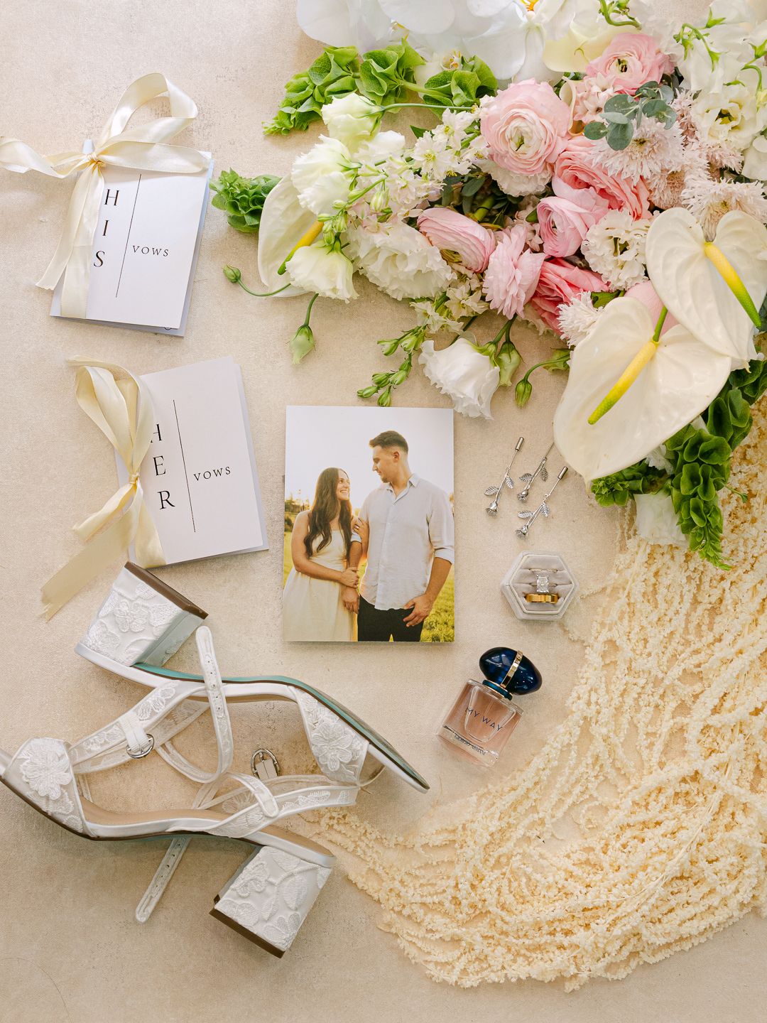 Elegant wedding flat lay with vow books, bridal heels, bouquet, perfume, and couple photo on neutral background.