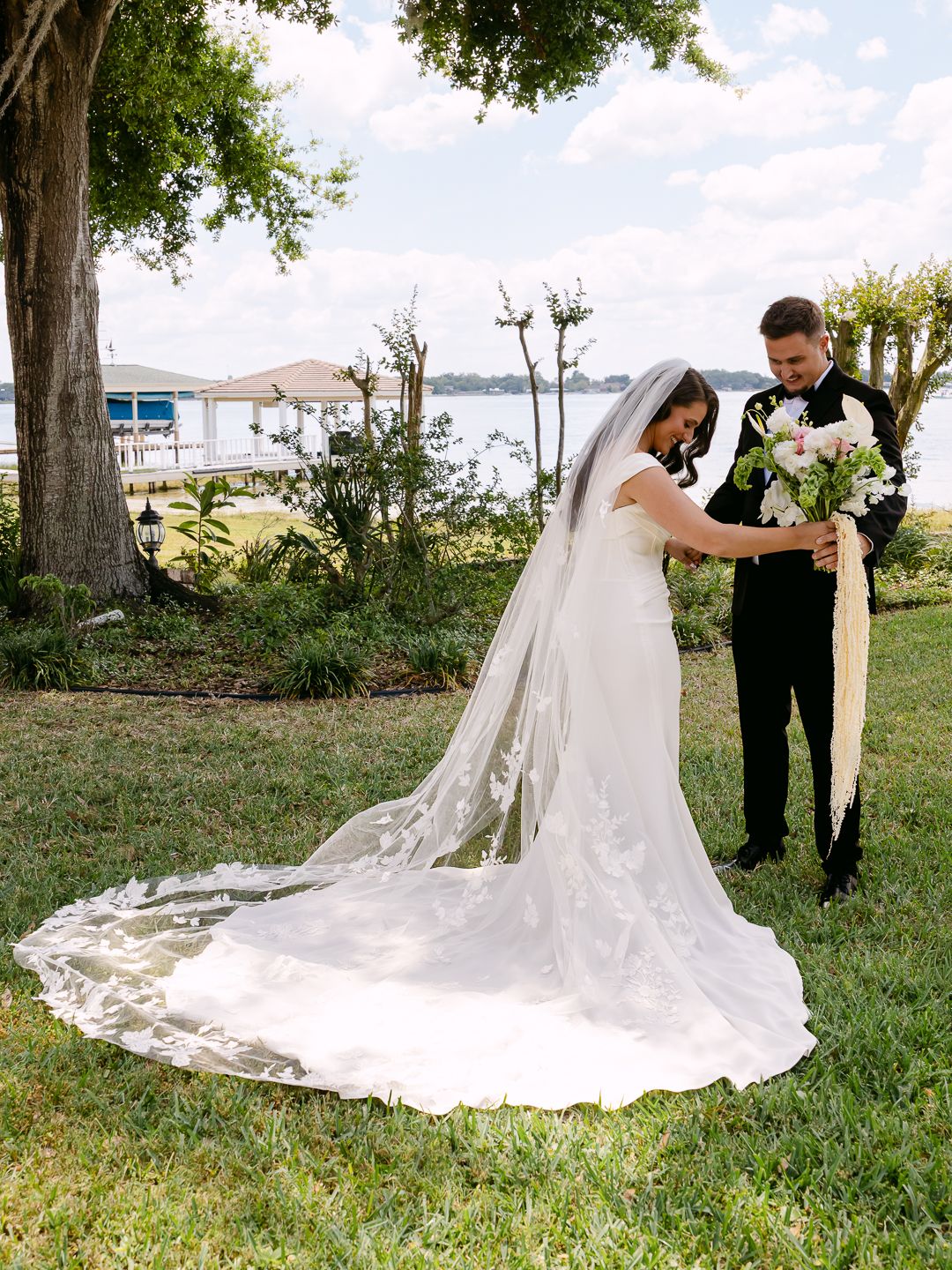 Bride and groom share a tender moment on a lakeside lawn, her cathedral veil trailing elegantly on the grass.