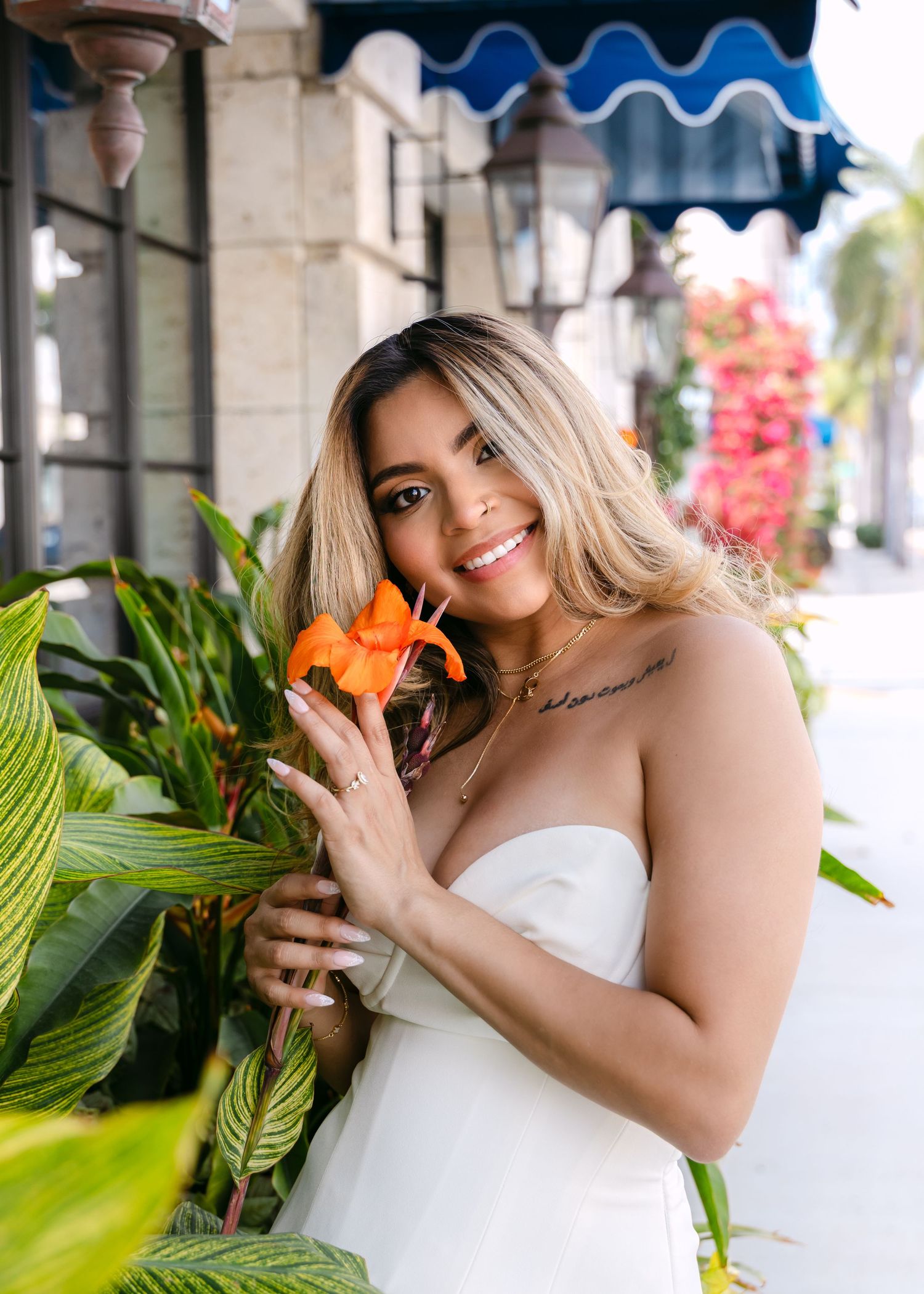 Smiling woman in white strapless dress holding an orange lily flower, posing outdoors near tropical plants.
