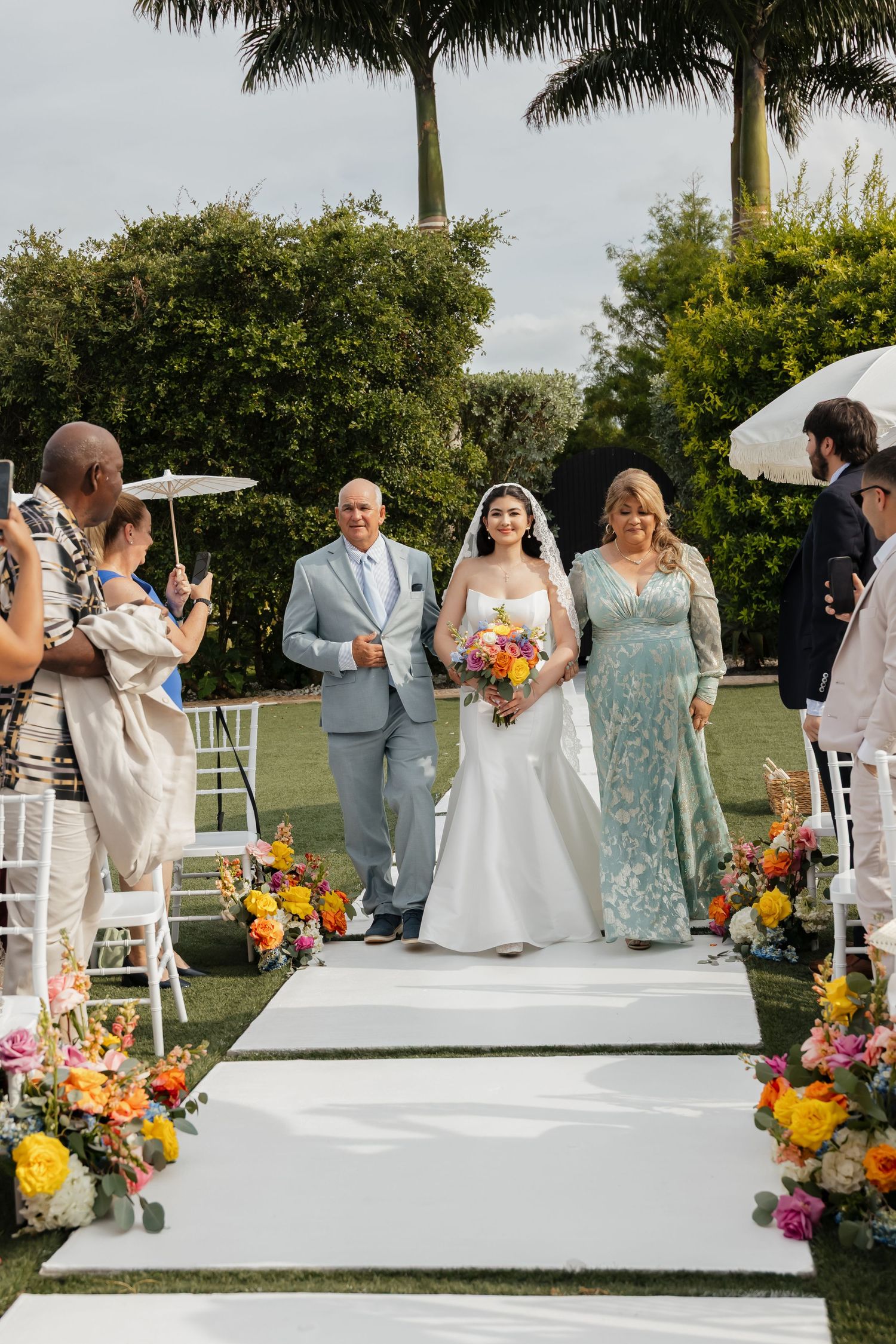 Bride in white gown and veil walking down the aisle with parents at an outdoor garden wedding.