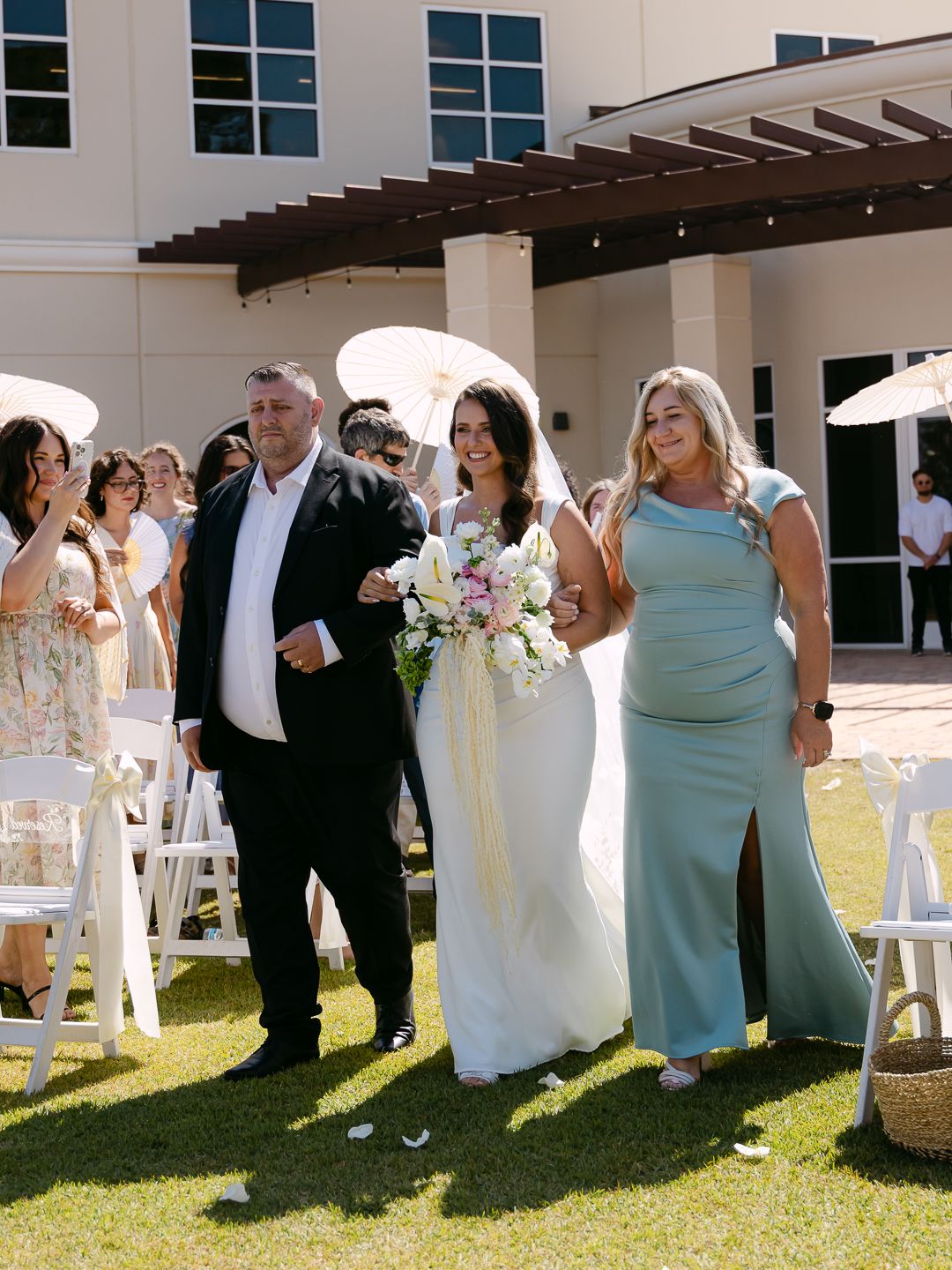 Bride walking down the aisle escorted by parents at sunny outdoor wedding ceremony with white chairs.