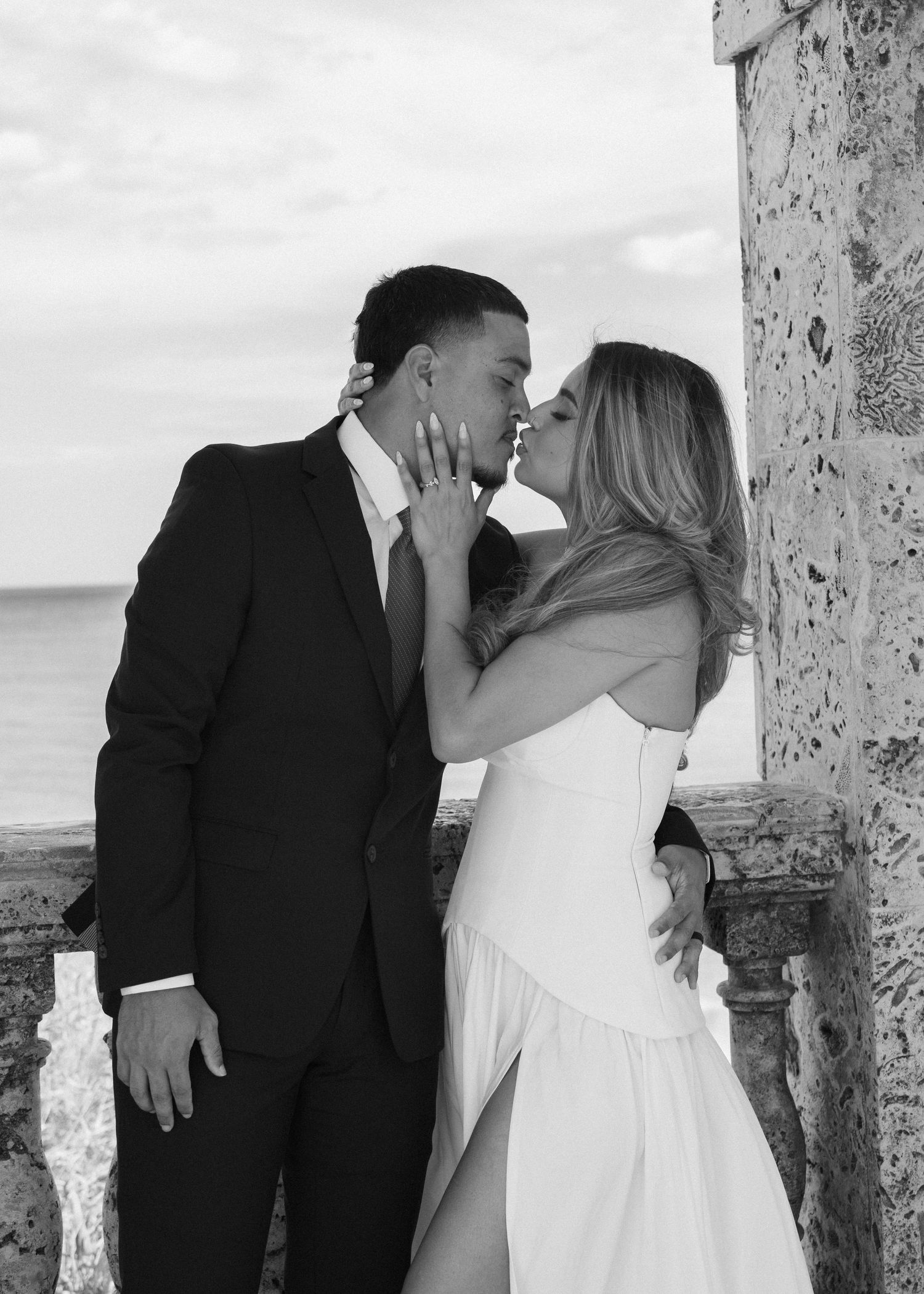 Black and white photo of a couple kissing, man in suit and woman in white dress, with ocean view backdrop.