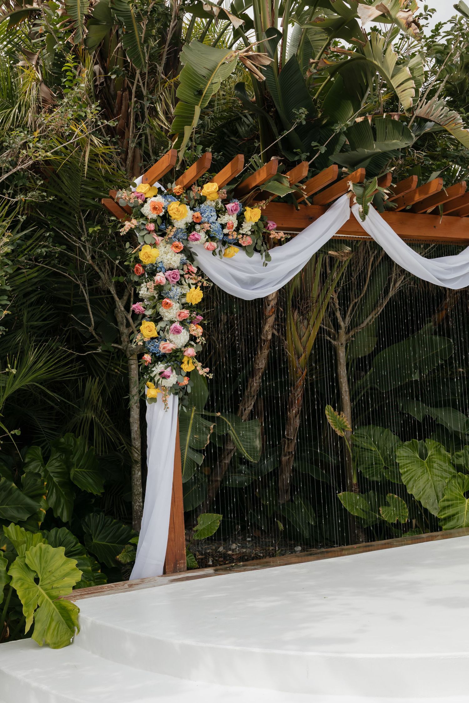 Elegant outdoor wedding arch adorned with colorful flowers and white draping amid lush tropical greenery.