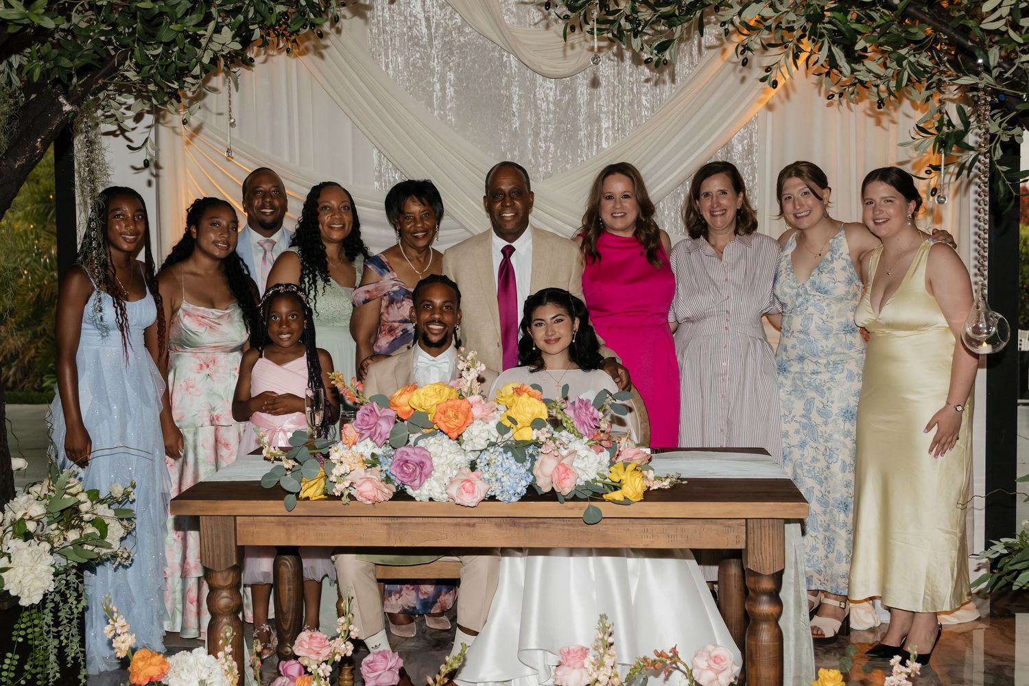 Wedding party group photo with bride and groom signing documents at a decorated table.