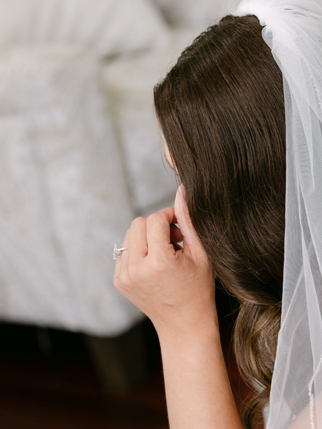 Bride with dark wavy hair adjusts her earring while wearing a delicate wedding veil.