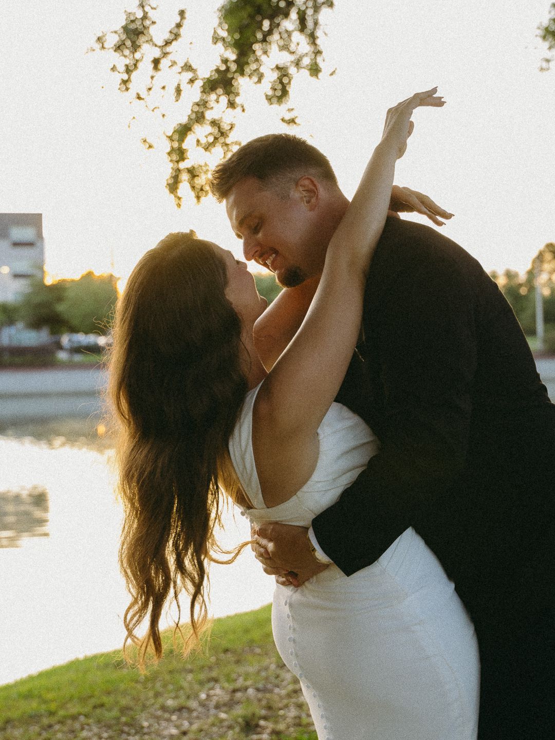 Couple sharing a romantic embrace at sunset near a lake, woman in white dress with long dark hair, man in black suit.