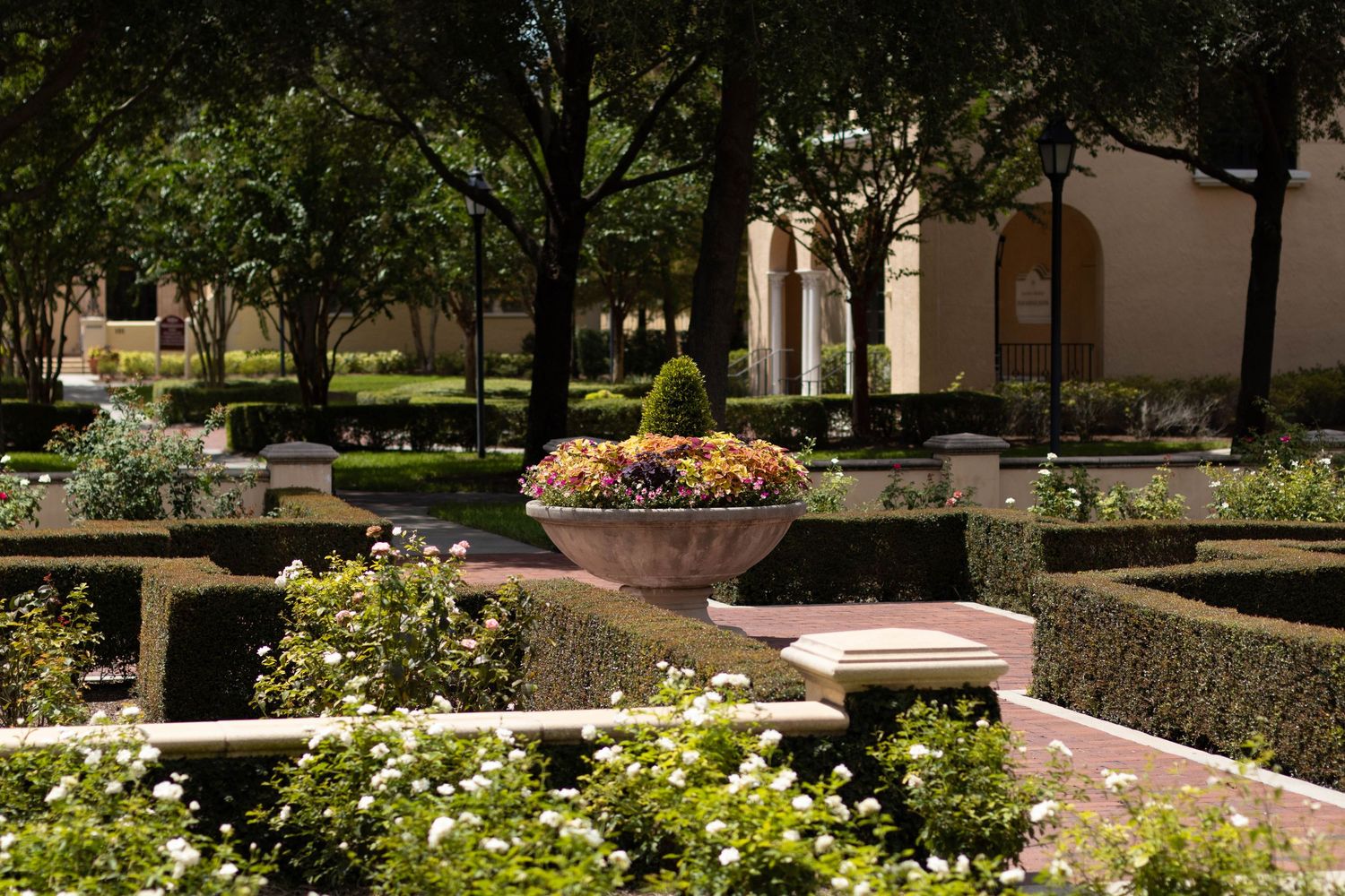 Formal garden with manicured hedges, colorful flower urn centerpiece, and shaded trees surrounding an elegant outdoor space.