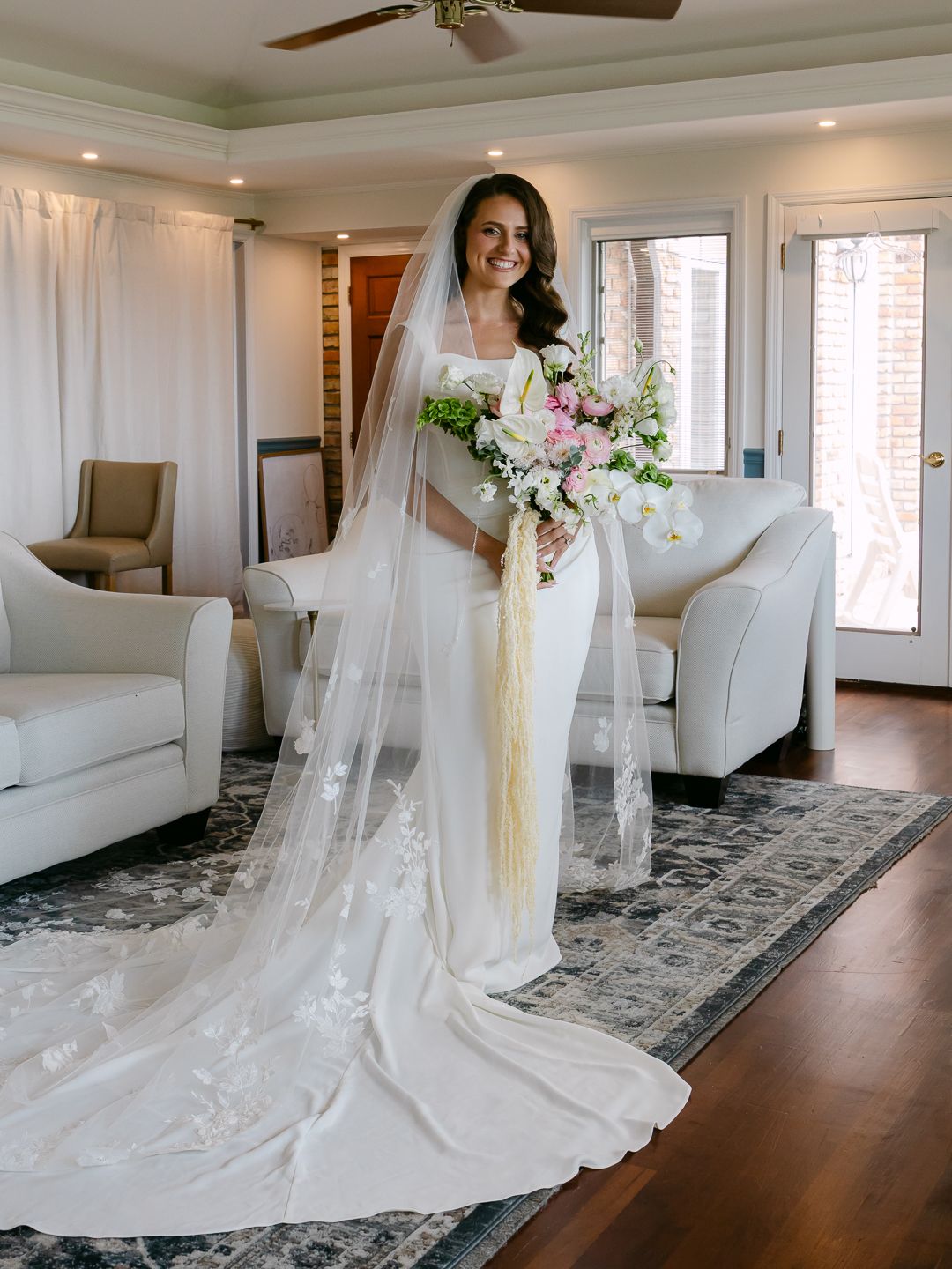 Bride in white mermaid gown with lace cathedral veil holds pink floral bouquet indoors.