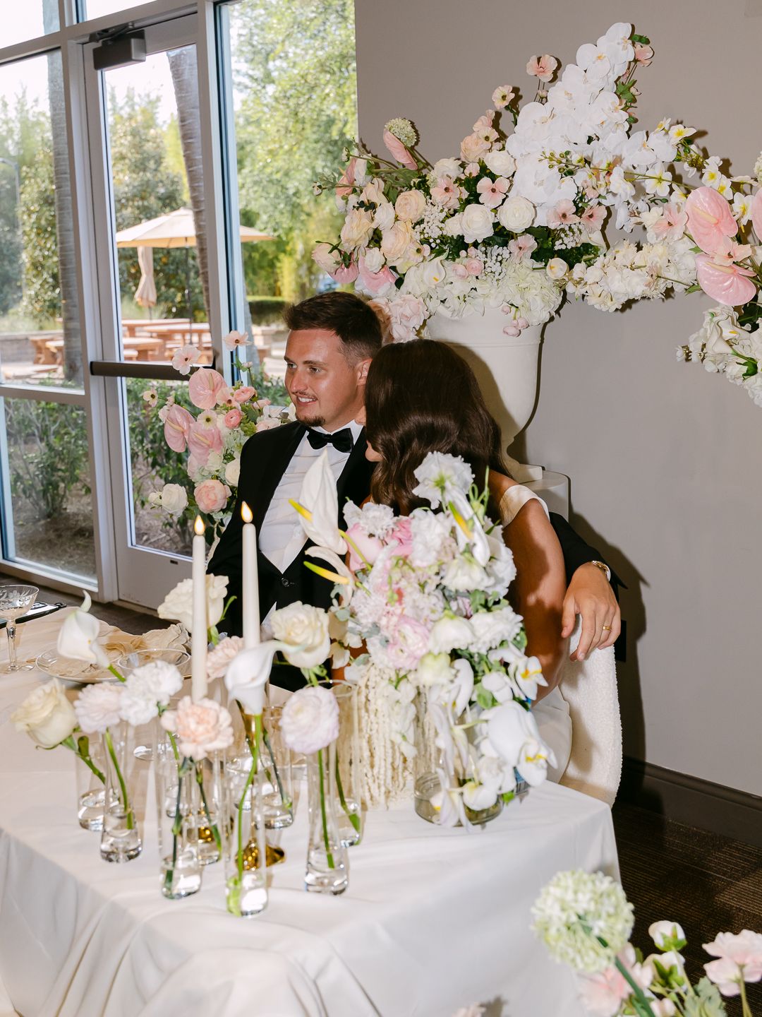 Bride and groom embrace at sweetheart table surrounded by lush white and pink floral arrangements.