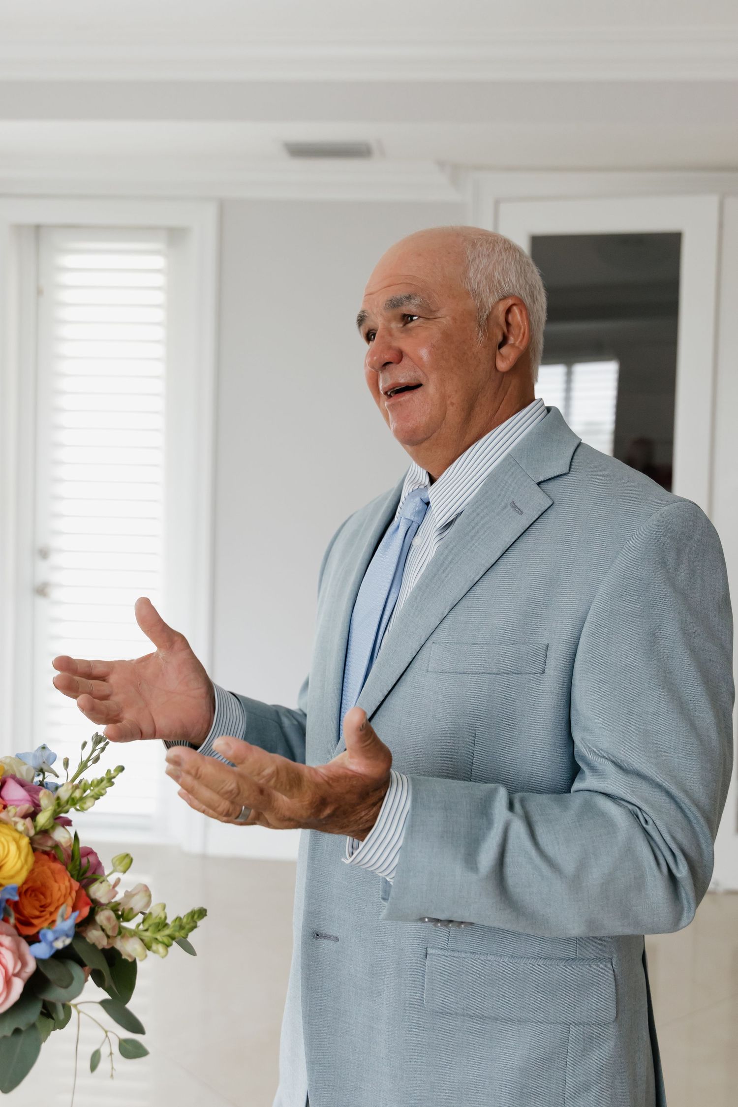 Elderly man in light gray suit gesturing while speaking at a wedding ceremony indoors.
