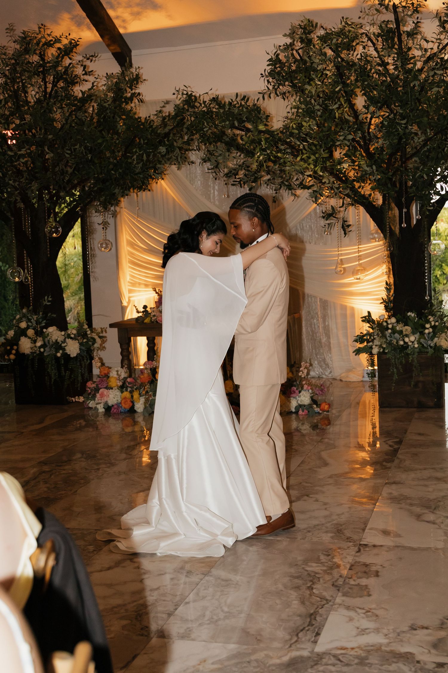Bride and groom share first dance under glowing trees with floral arrangements at elegant indoor reception.
