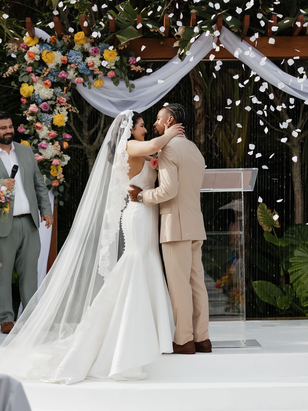 Bride and groom share first kiss at outdoor wedding ceremony with colorful floral arch and white rose petals falling.