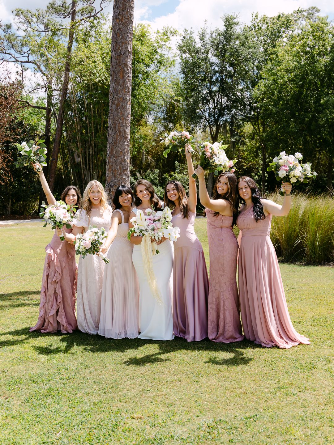 Bride and six bridesmaids in pink dresses celebrate outdoors raising floral bouquets in sunny garden.