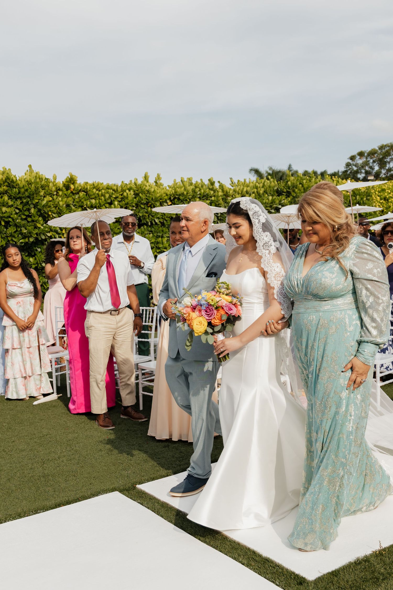 Bride walks down outdoor aisle with parents, holding colorful bouquet, wearing white gown and lace veil.