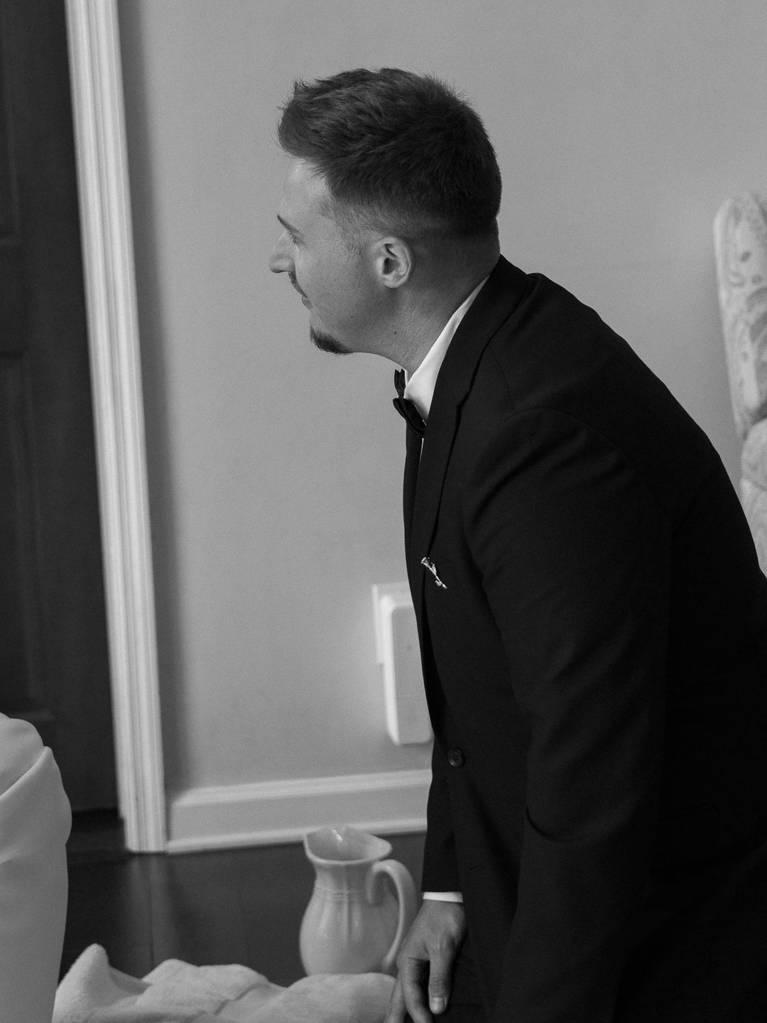 Black and white portrait of groom in tuxedo with bow tie seen from the side in a softly lit room.