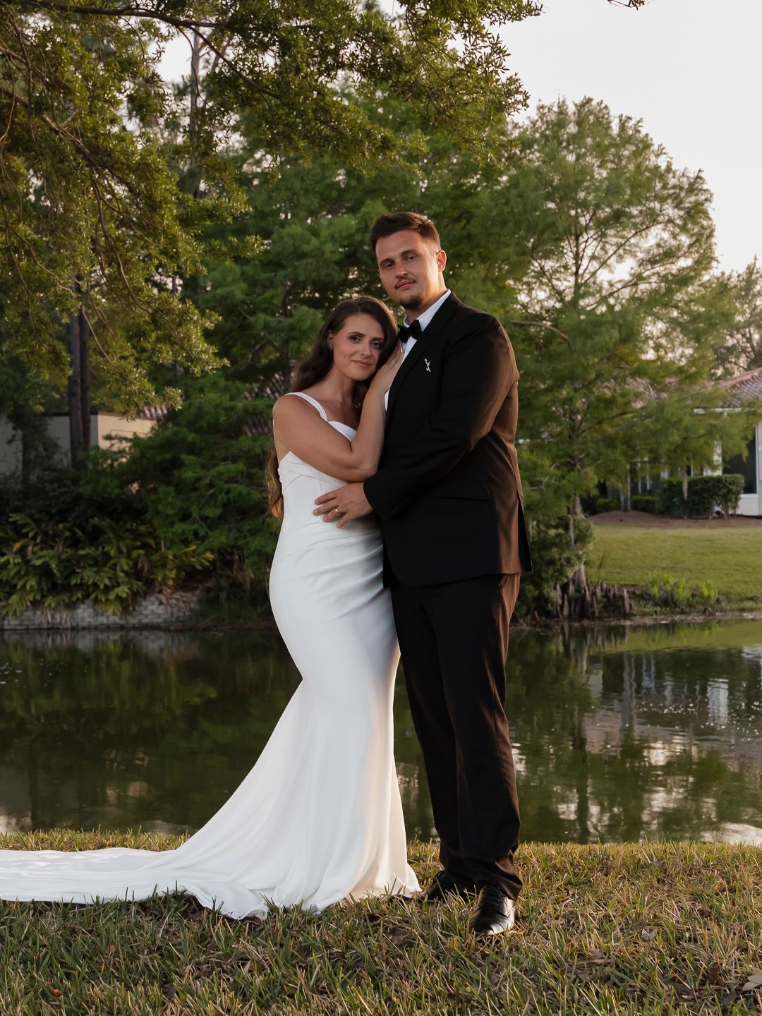 Bride and groom pose elegantly by a pond at golden hour in wedding attire.
