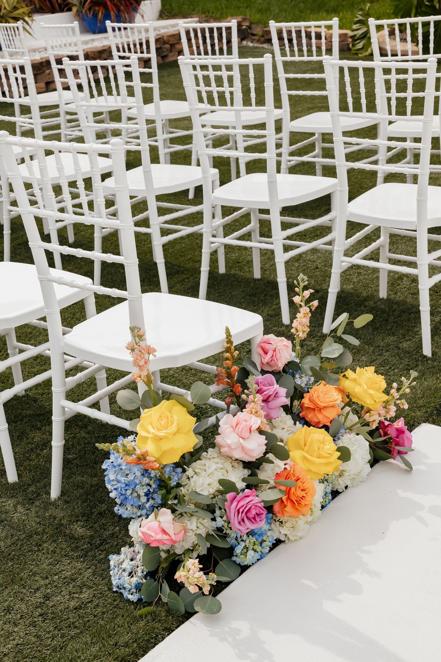 Colorful floral arrangement with yellow, pink, and orange roses lining a wedding ceremony aisle with white Chiavari chairs.