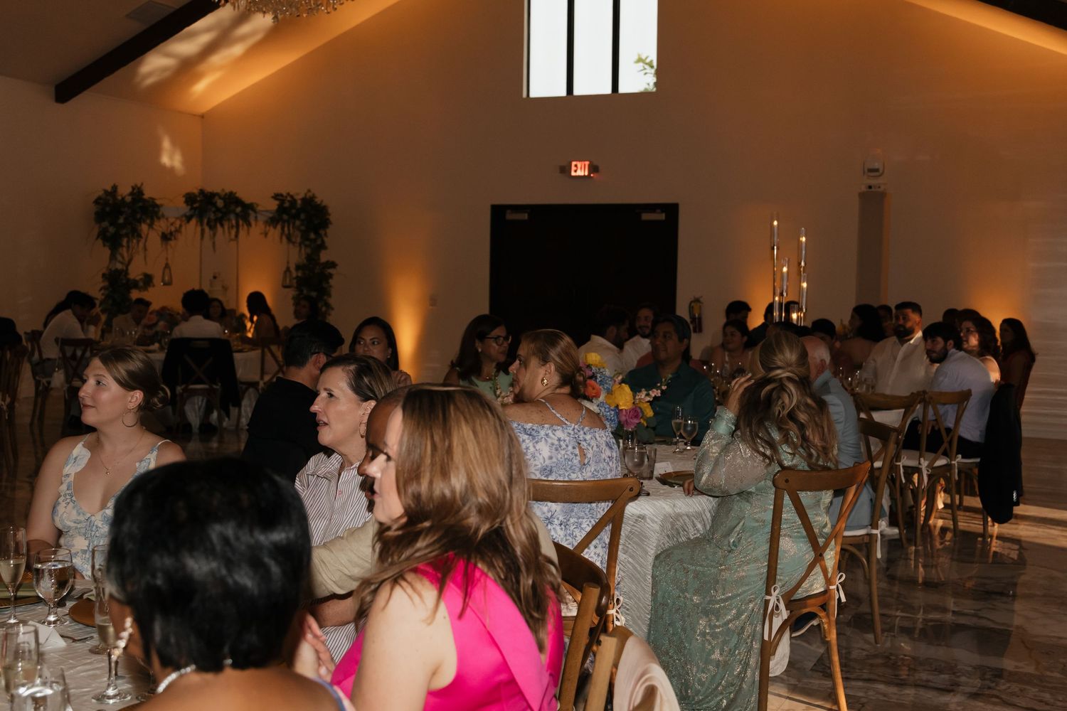 Elegantly dressed wedding reception guests seated at tables in a warmly lit white reception hall.