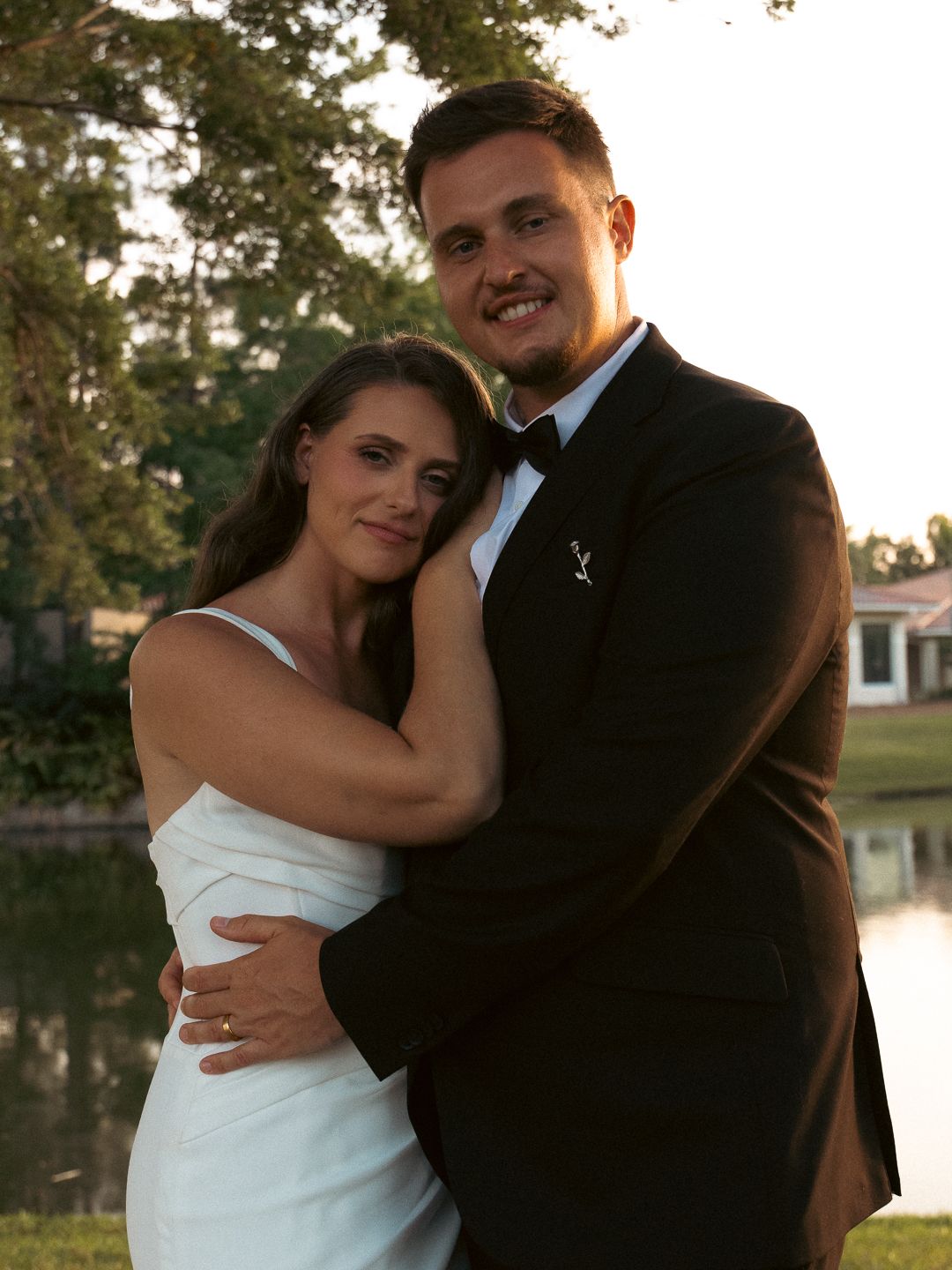 Couple in formal attire embracing outdoors near a pond, woman in white dress and man in black tuxedo.