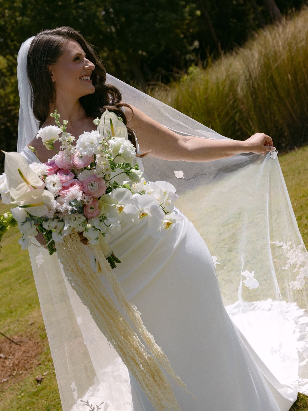 Smiling bride in white gown holding large pink and white floral bouquet while spreading her veil outdoors.