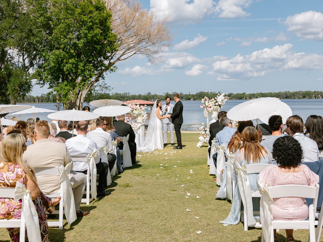 Outdoor lakeside wedding ceremony with guests holding white parasols as couple exchanges vows at floral arch.