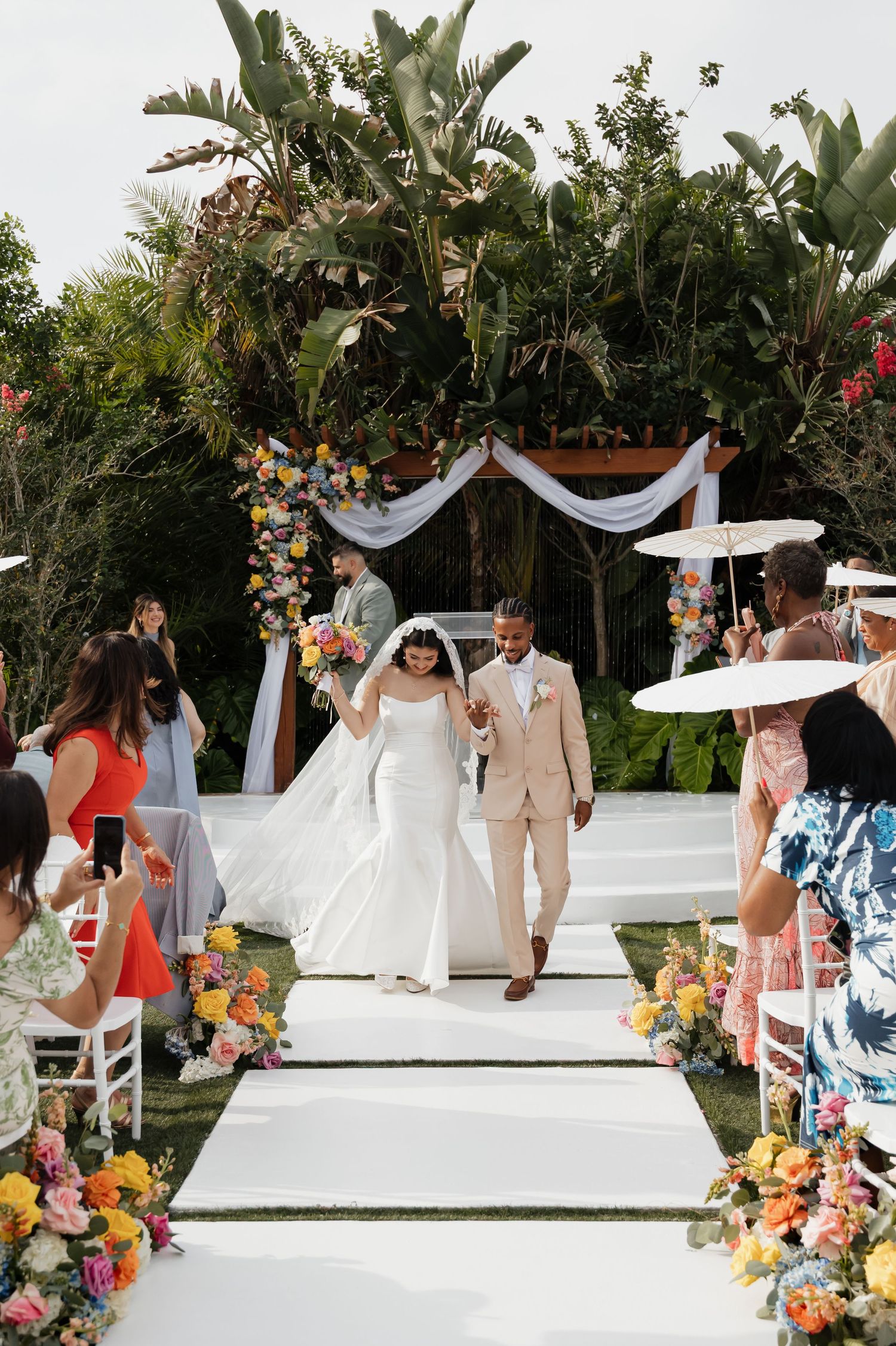 Bride and groom walking down white aisle after wedding ceremony surrounded by colorful floral arrangements.