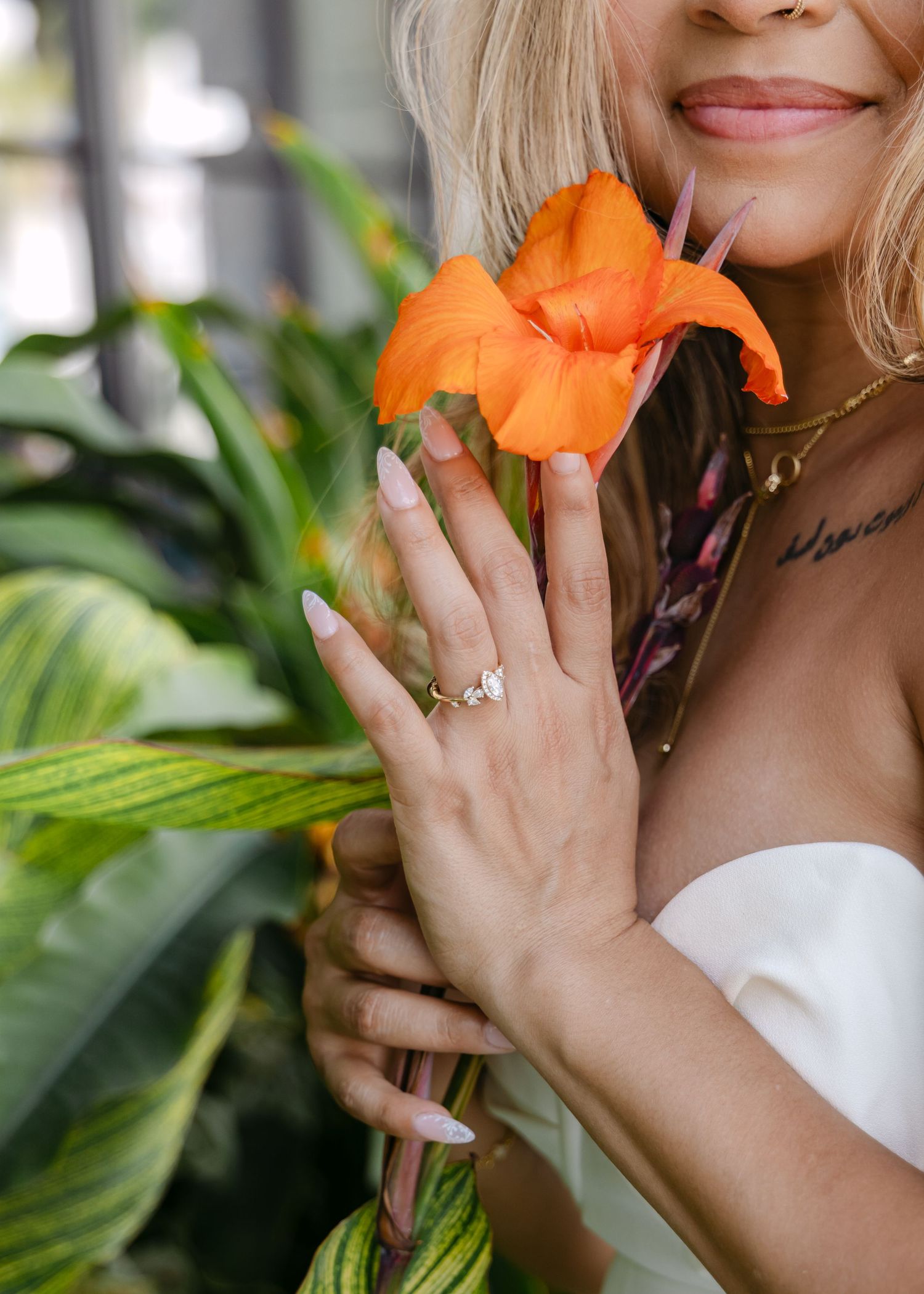 Smiling bride holding an orange flower, showcasing a diamond engagement ring, with lush green tropical plants in the background.
