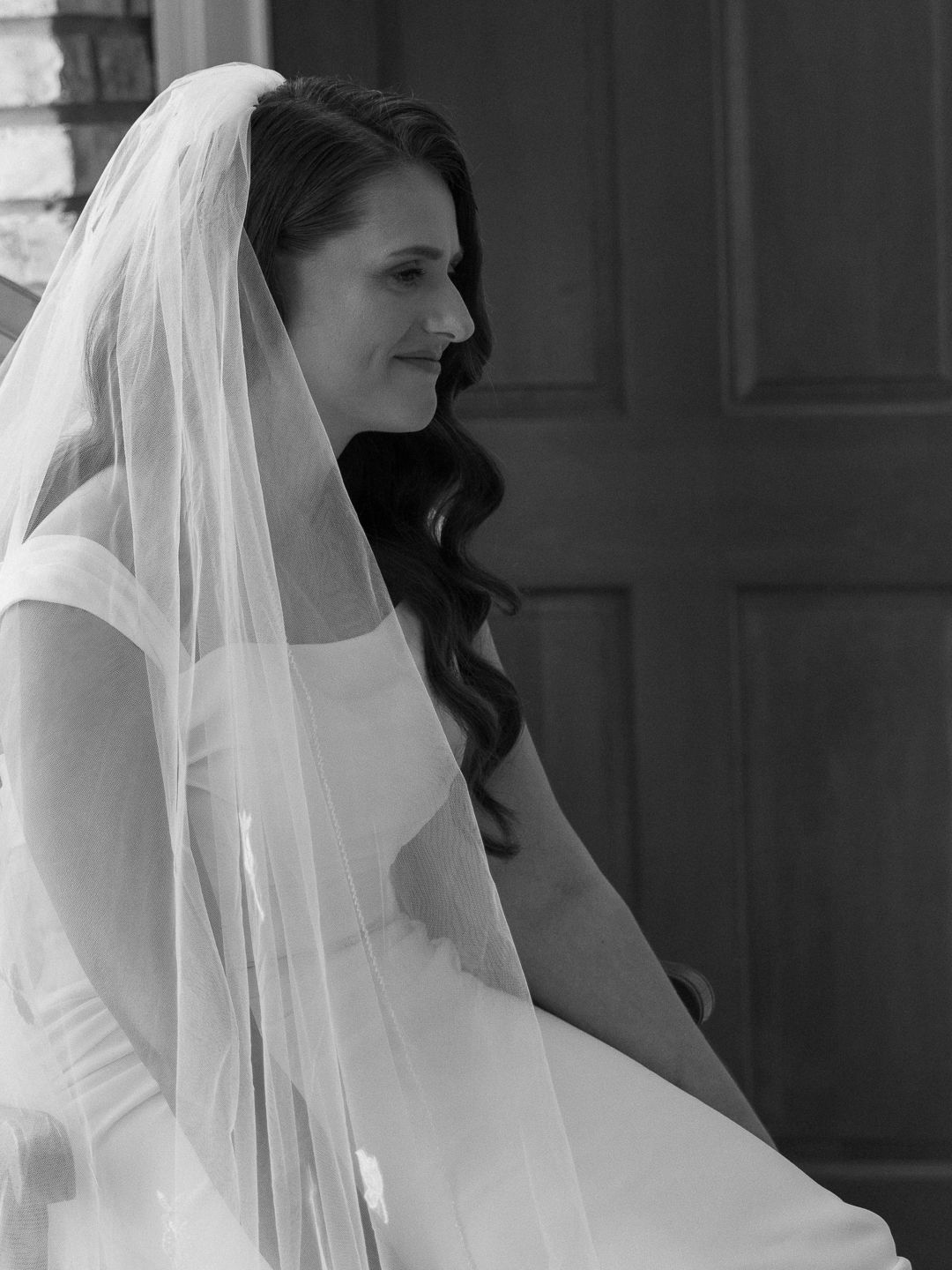 Black and white photo of a smiling bride in a white veil and gown seated near a wooden door.