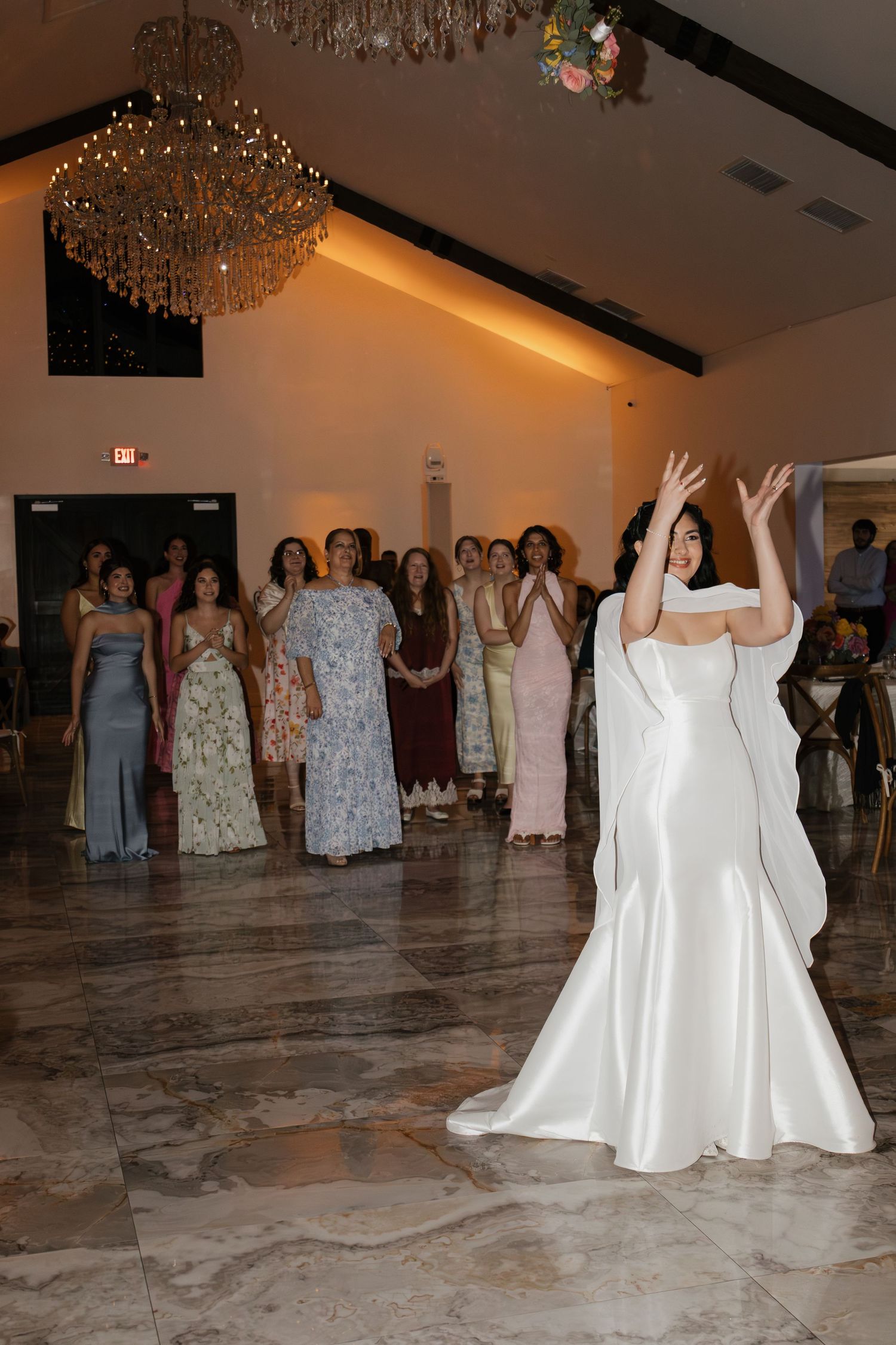 Bride in white gown tossing bouquet to group of women at wedding reception under chandelier.