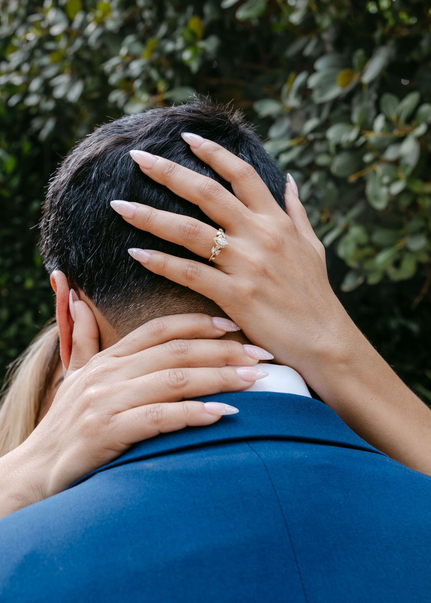 Bride placing her hand with a gold engagement ring on the back of her grooms neck outdoors.