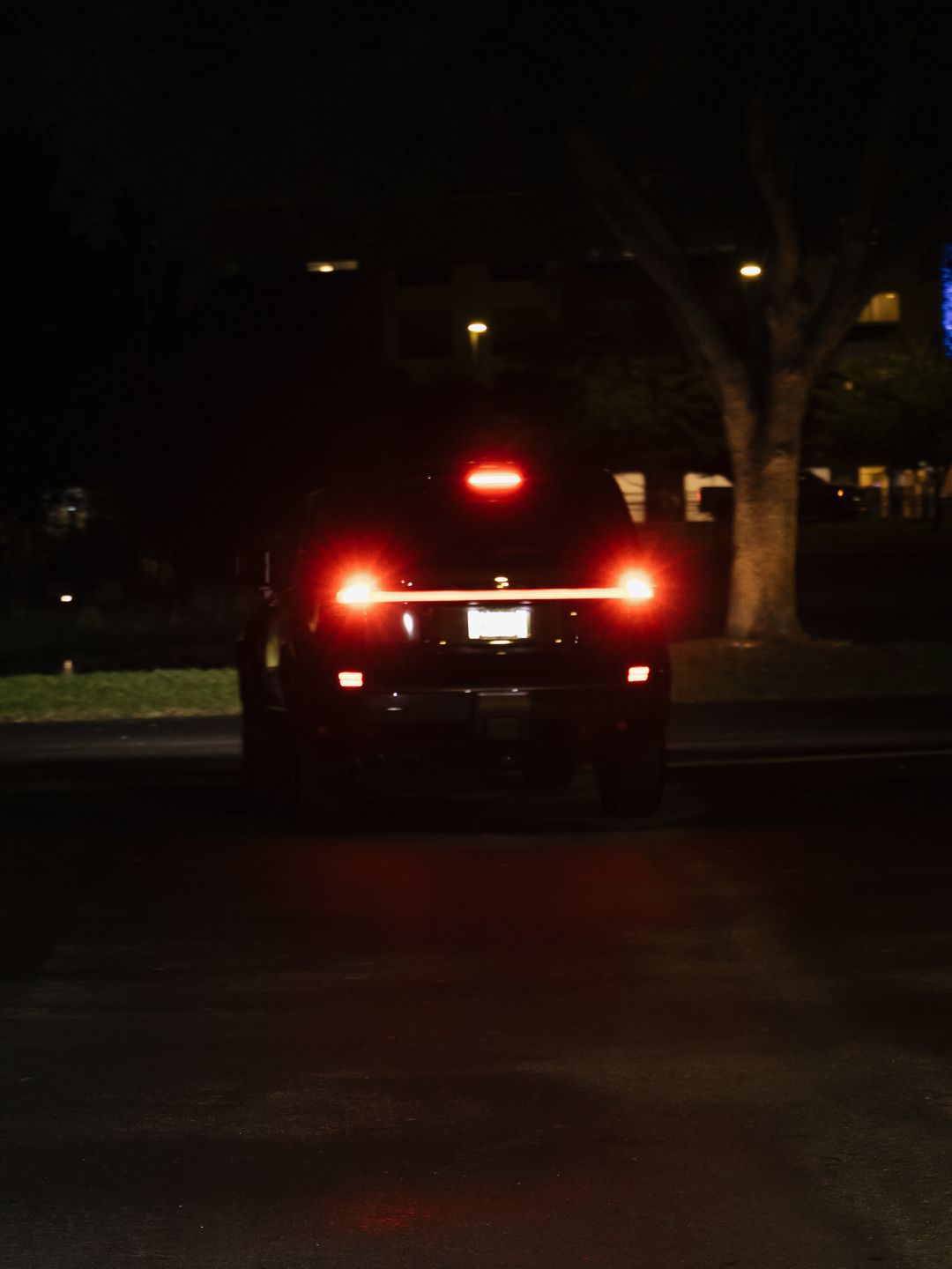 Dark nighttime photo of a large black SUV or truck from behind with red brake lights illuminated in a parking lot.