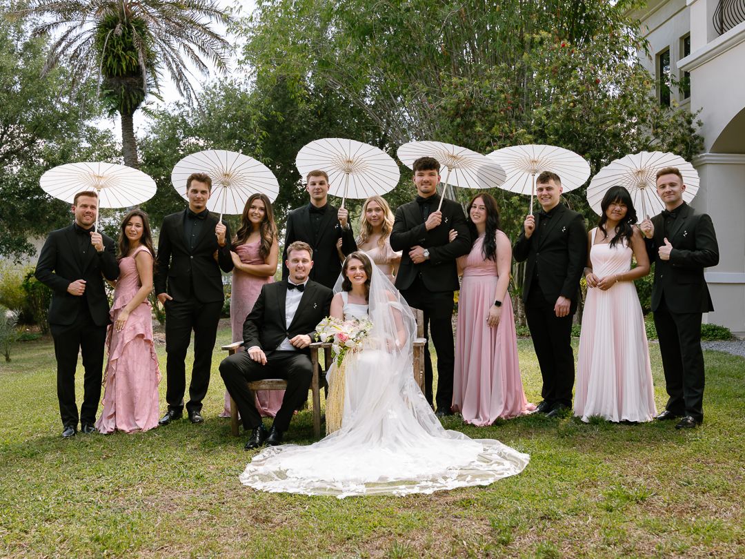 Wedding party in black suits and pink gowns holding white parasols pose with bride and groom on green lawn.