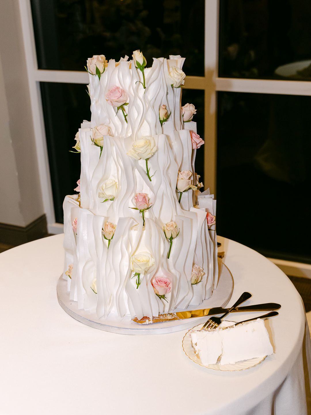 Three-tier white wedding cake with wavy texture and pink and cream roses on a white table.