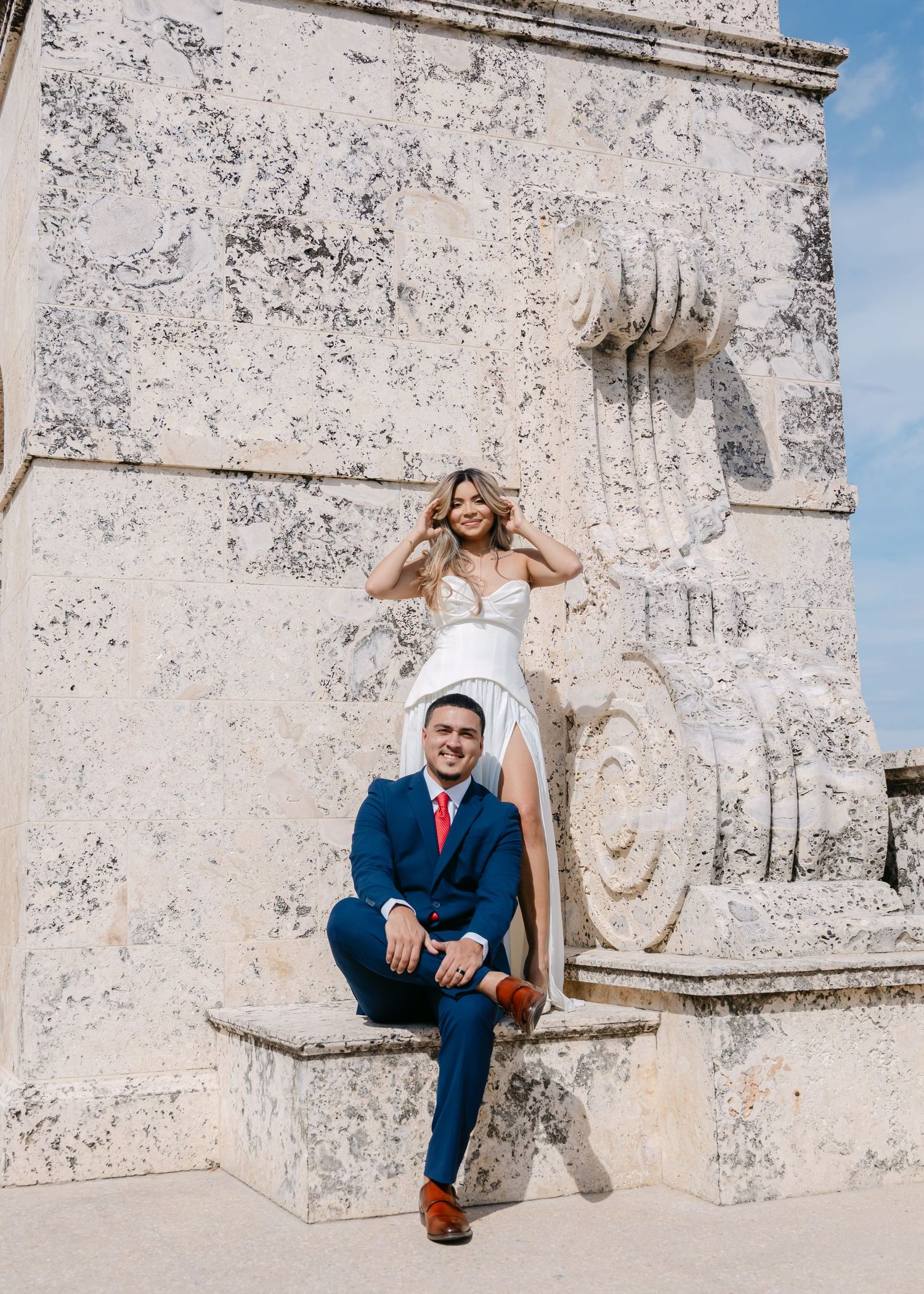 Bride in white gown and groom in blue suit pose on ornate stone monument outdoors.
