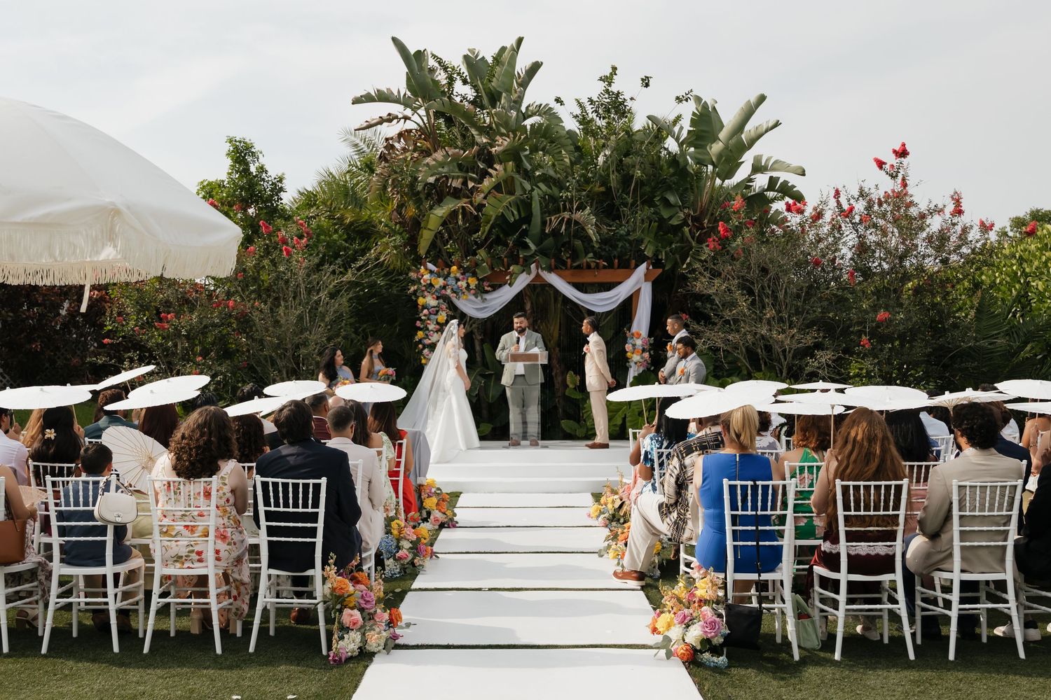 Outdoor wedding ceremony with guests seated in white chairs under umbrellas, couple at floral altar on white steps.