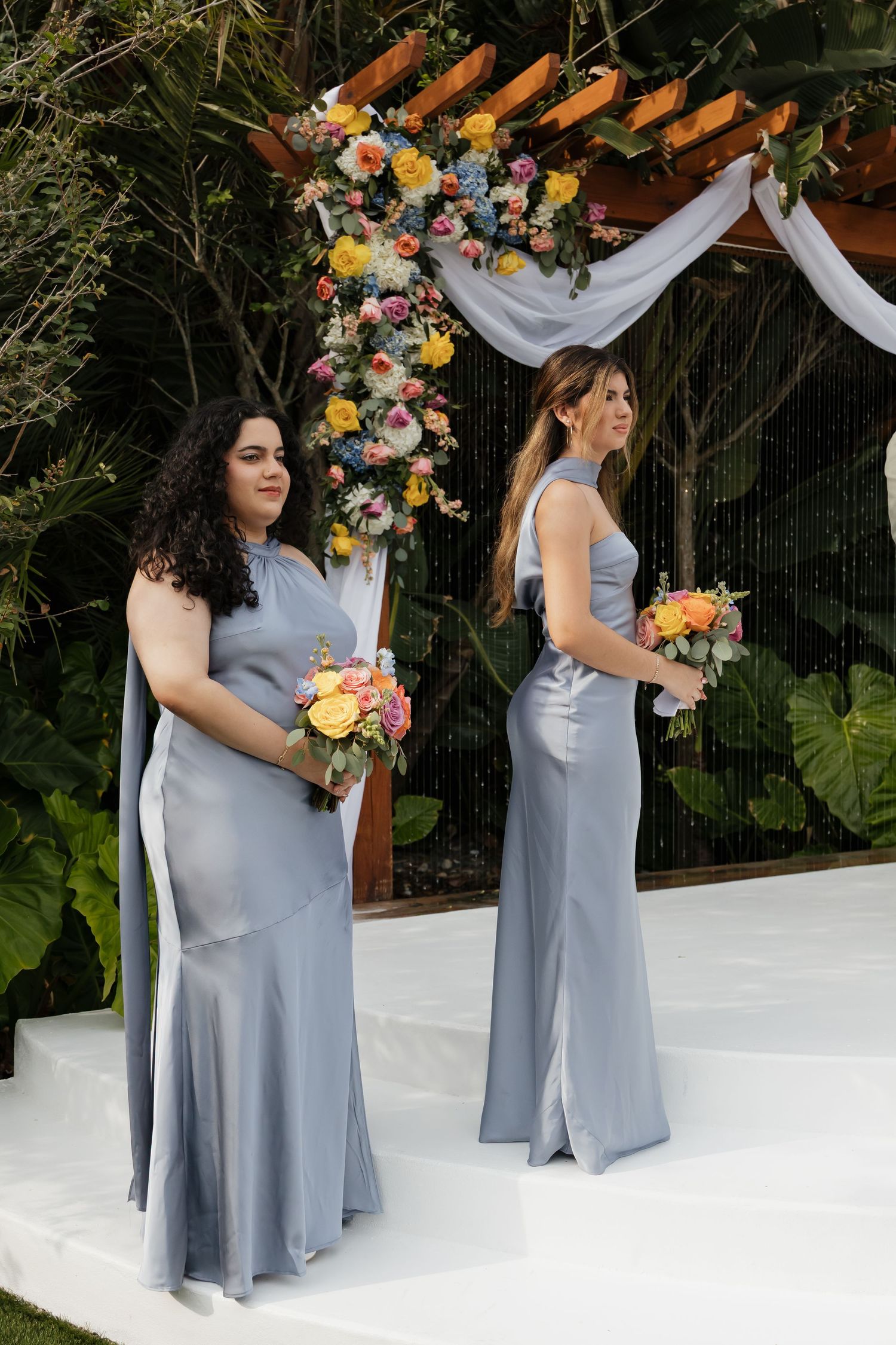 Two bridesmaids in dusty blue satin gowns holding colorful bouquets standing at a floral-decorated wedding altar.