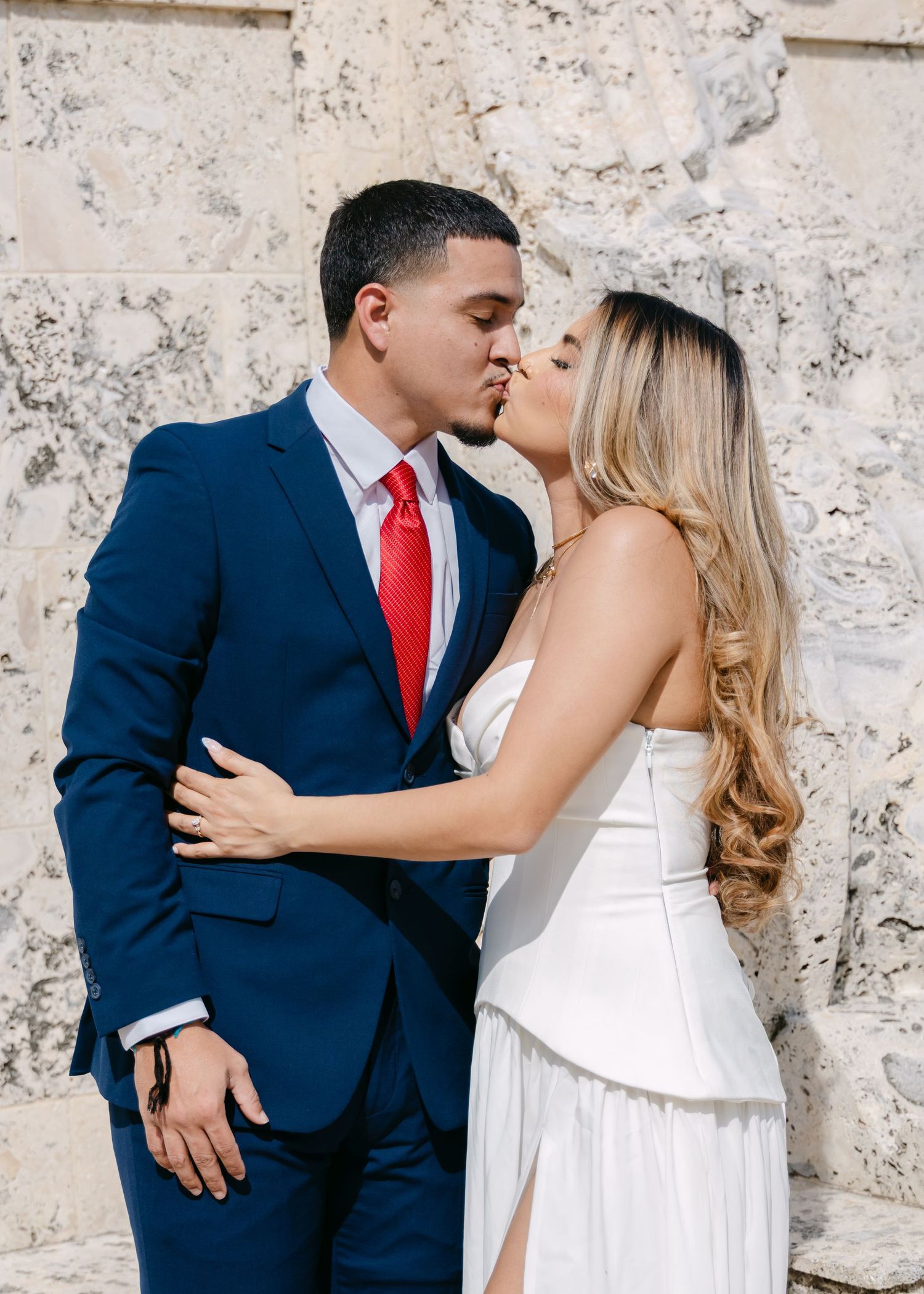 A couple shares a romantic kiss, the man in a navy suit with red tie and the woman in a white dress, against a stone wall.