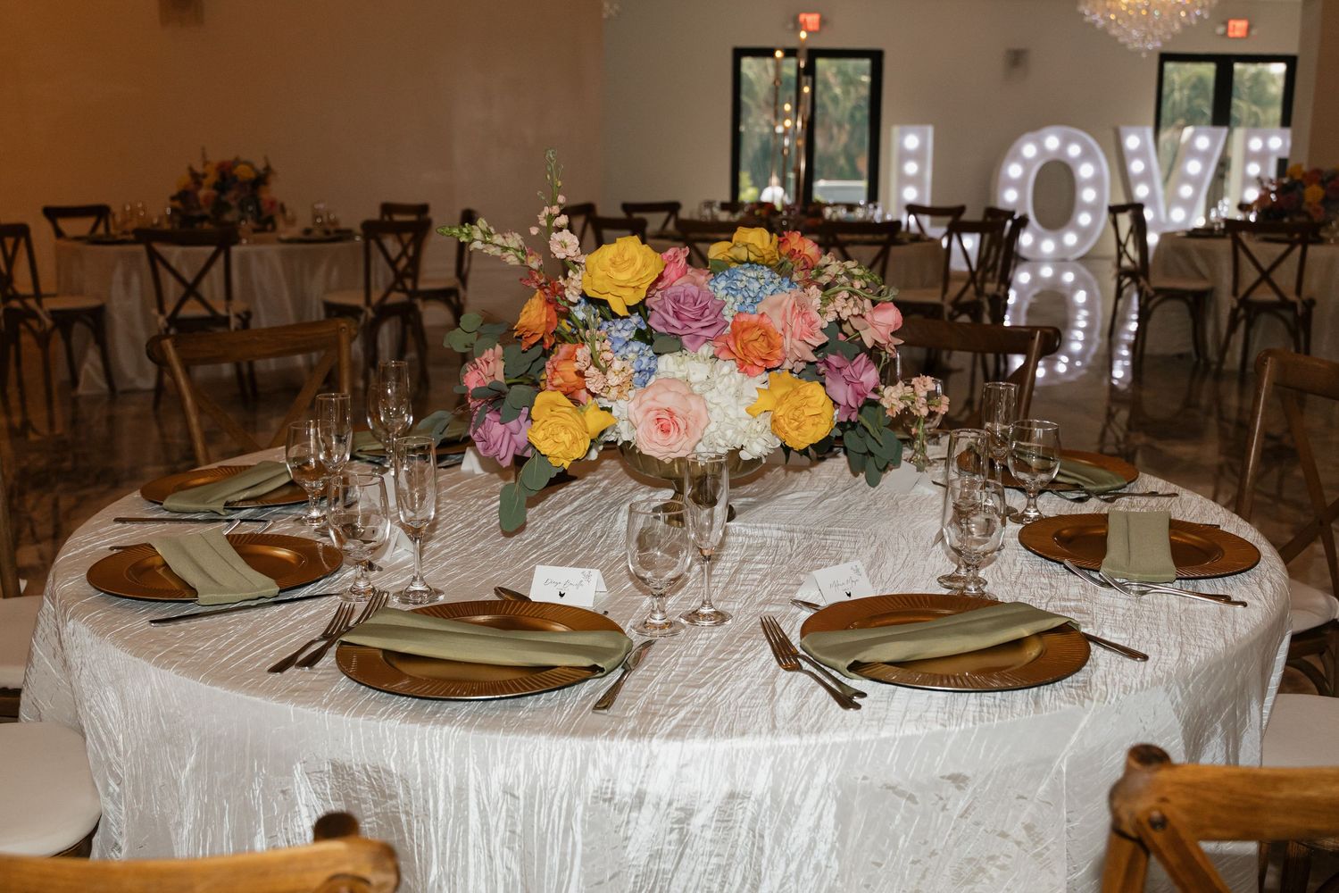 Round wedding reception table set with gold chargers, glassware, and a vibrant floral centerpiece.