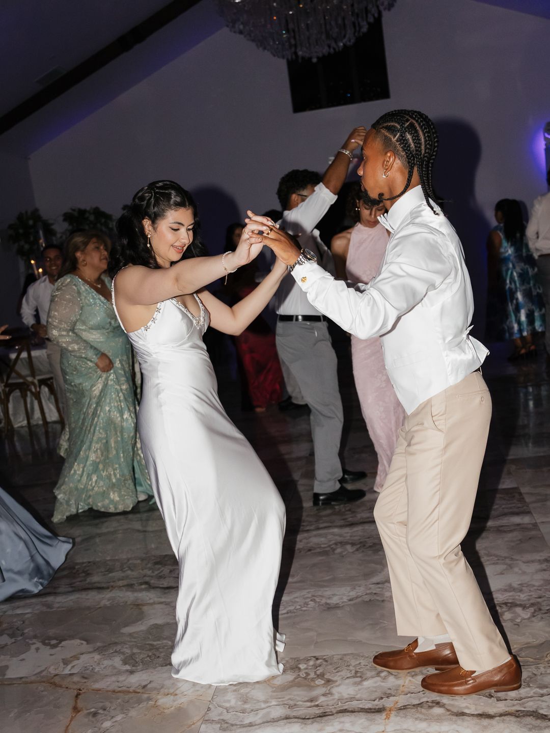Bride in white satin gown dances energetically with groom in white shirt at wedding reception.