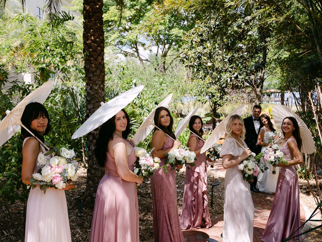 Bridesmaids in pink dresses holding bouquets and white parasols in a garden.