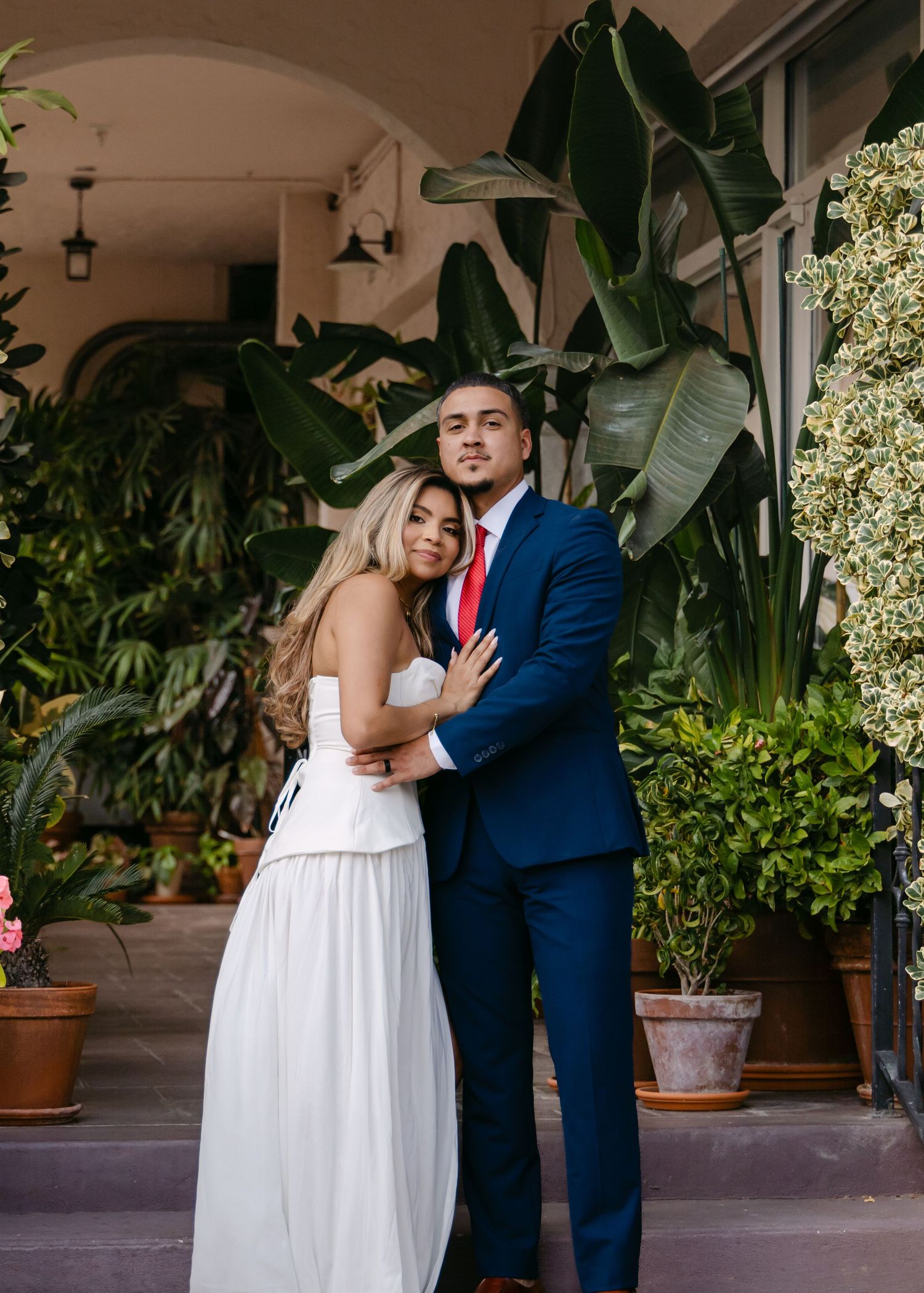 Couple posing together, bride in white dress and groom in navy suit with red tie, surrounded by lush greenery.