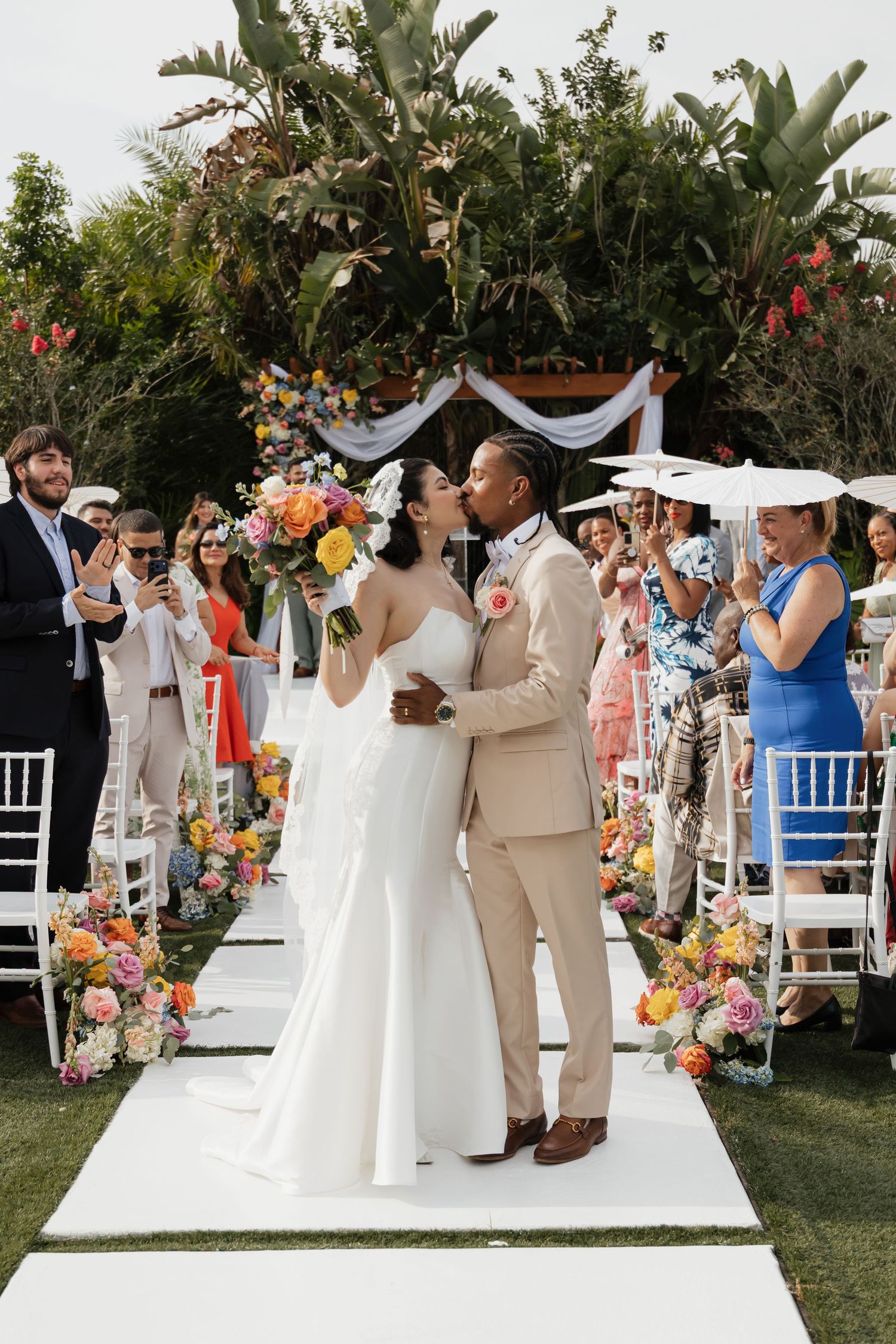 Newlyweds kiss on white aisle as guests celebrate at vibrant outdoor garden wedding ceremony.