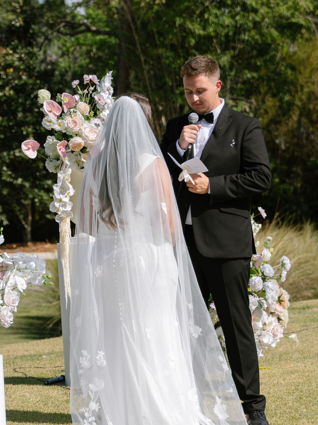 Groom in black tuxedo reads vows to bride at floral arch during outdoor wedding ceremony.