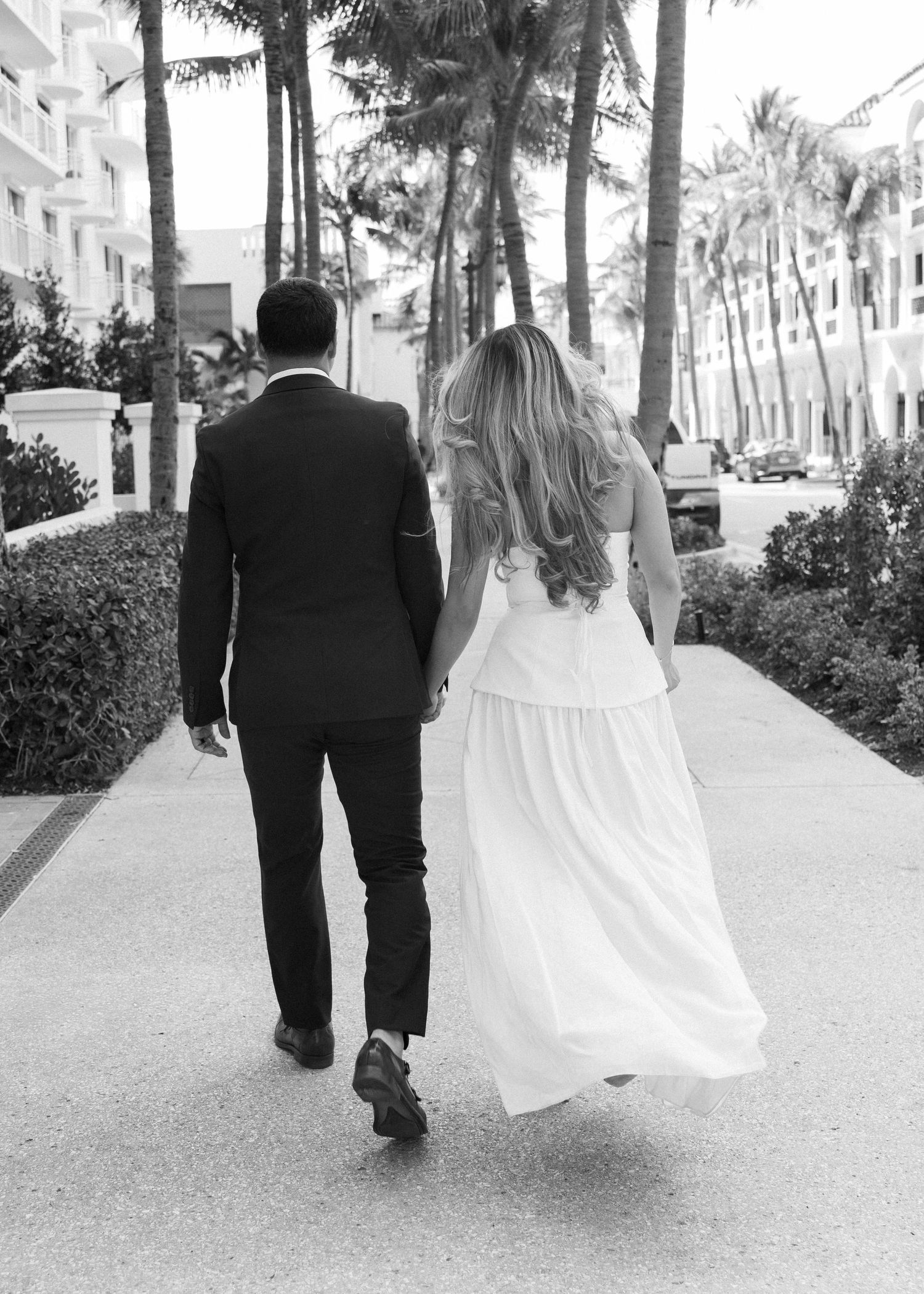 Black and white photo of a bride and groom walking hand in hand along a palm tree-lined path.