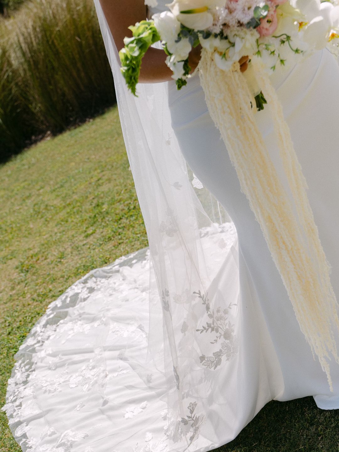 Bride in a white wedding gown with lace cathedral train holding a bouquet of white and pink flowers outdoors.