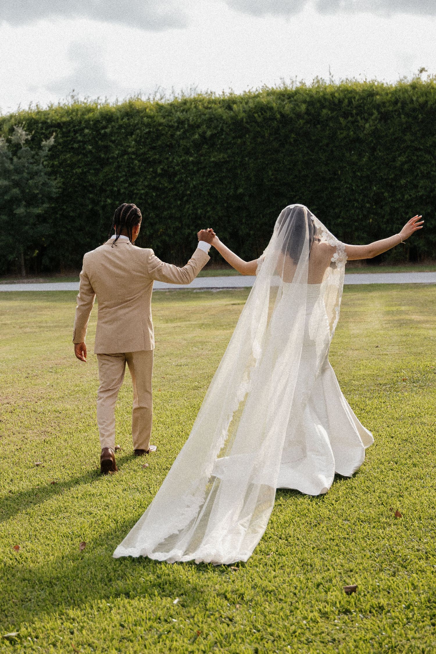 Bride in white gown and veil holds groom's hand in beige suit as they walk across lush green lawn.